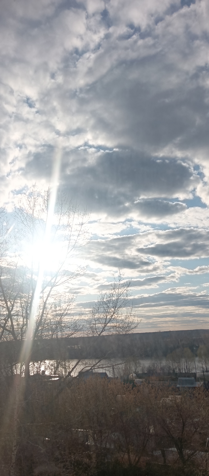 The image depicts a landscape with the sky occupying the majority of the frame. Clouds are scattered across the sky, varying in color from bright white to dark gray. A bright light source, potentially the sun, is positioned behind a tree on the left side, creating a strong glare and obscuring the view in that area. The tree is bare, with leafless branches silhouetted against the bright light.
In the middle ground, there appears to be a body of water, possibly a river or lake. The far shore is visible, featuring a line of trees and some structures that may be buildings or houses.
The foreground consists of more trees and bushes, appearing somewhat blurry and out of focus. The overall scene has a muted color palette, with shades of gray, white, and brown dominating.
The location within the city of Unknown, Unknown is not determinable from the image.