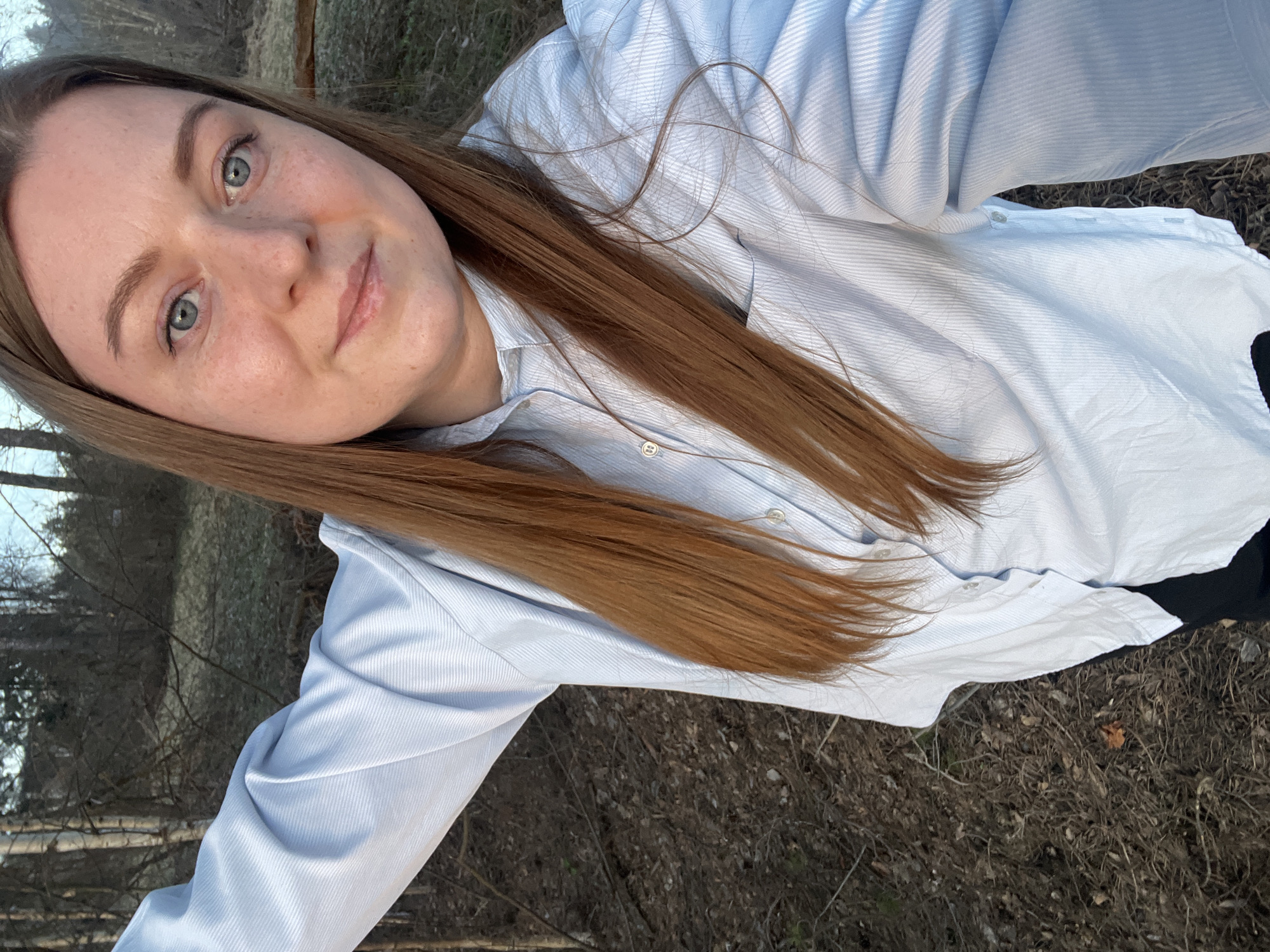 The image shows a woman with auburn hair and light-colored eyes. She is wearing a light blue button-down shirt. The background appears to be a natural setting with trees and dry vegetation, suggesting an outdoor location in Раздолье, Russia. The photograph is taken at an angle, possibly a selfie.
