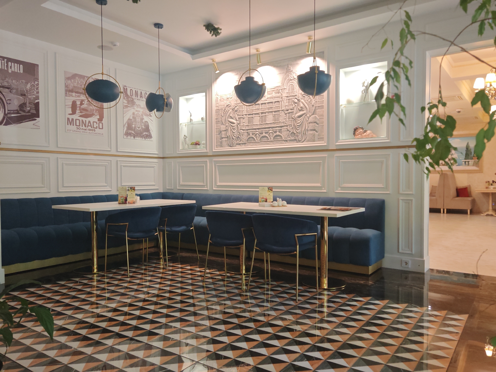 This image captures the interior of an elegantly decorated cafe or restaurant, characterized by a blend of classic and modern design elements. The scene features two long, white rectangular tables with golden bases, surrounded by plush blue velvet chairs with gold-colored frames. Along the wall, there's a continuous banquette seat upholstered in matching blue velvet.
The walls are predominantly white, adorned with decorative molding in a wainscoting style and accented with thin gold trim. On the left wall, three framed posters depict vintage car racing scenes, two prominently featuring "MONACO" and one "MONTE CARLO." Visible text includes "30 mai 1965", "23° GRAND PRIX AUTOMOBILE", "27 MAY 2012", and a "ups" logo. Above the banquette, a large, ornate white bas-relief sculpture depicts an architectural facade flanked by two figures, possibly angels. Built-in display niches with glass shelves showcase various decorative objects, including a small, cherub-like statue.
From the ceiling, several distinctive pendant lights hang, featuring blue, bowl-shaped shades encircled by gold-colored rings. Recessed spotlights illuminate the wall art. The flooring is a striking geometric pattern composed of triangles in black, white, and a brownish-orange hue, with a highly reflective, polished surface. A leafy plant branch is visible in the foreground on the right, partially obscuring the view into an adjacent, brighter room with a different floor and furniture, including a chandelier.
The setting appears well-lit, suggesting daytime or a brightly illuminated interior. No people are present, indicating a quiet moment before or after service. The overall ambiance is sophisticated and inviting.