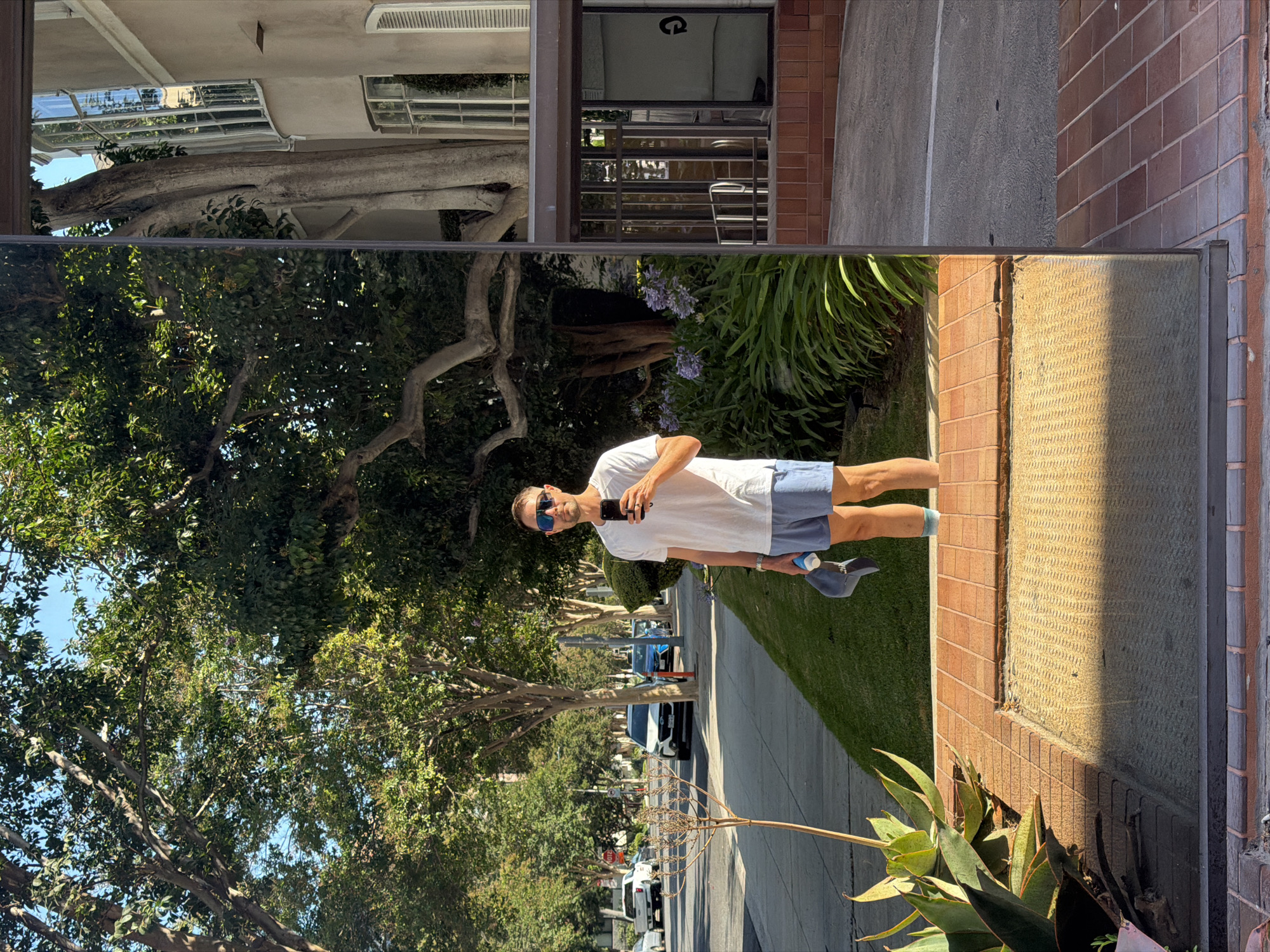 A man stands reflected in a glass surface in Beverly Hills, United States of America, holding a smartphone and a cap. He is wearing a white t-shirt, blue shorts, and blue socks. The reflection shows a street with cars and trees, and a building with windows. The man is positioned in front of lush green plants and a brick walkway. The sun casts a shadow on the walkway, indicating it is daytime.