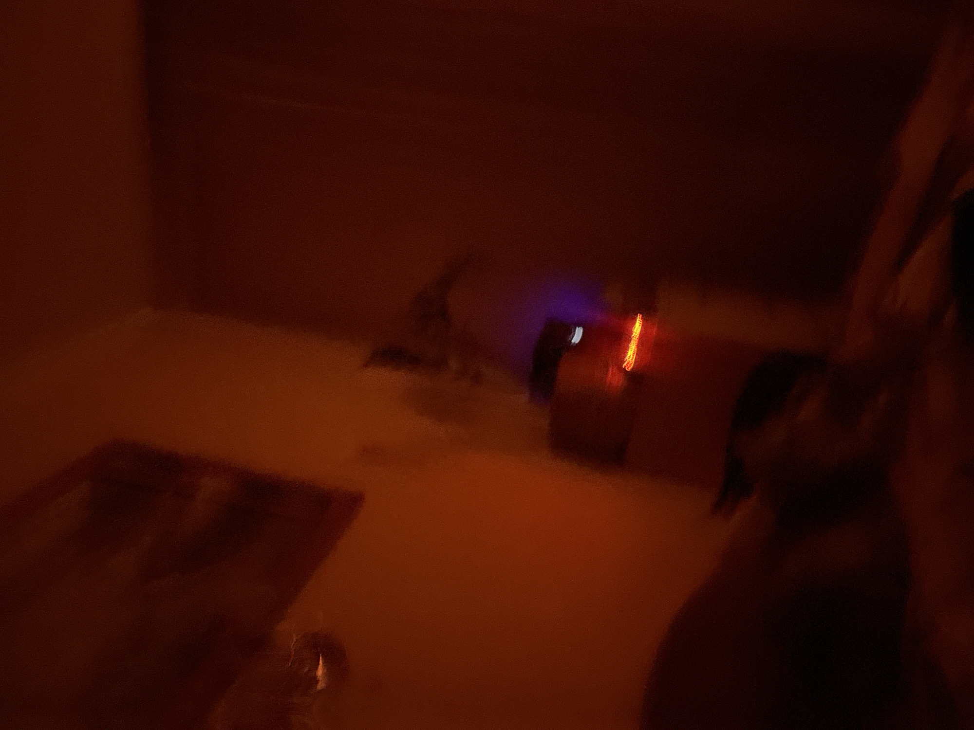 The image shows a dimly lit room with reddish-orange hues, suggesting it might be nighttime or have subdued lighting. The scene is heavily blurred, making it difficult to discern specific details. In the background, there appears to be a dark surface, possibly a wall or furniture, with some faint light sources visible, including what looks like a red or orange light streak and a purplish glow. The foreground is mostly dark and indistinct, with a vague shape that could be a person or an object. The overall impression is one of a private, possibly domestic, space. The provided metadata indicates the location as Kyiv, Ukraine, but there are no visual cues in the image that specifically confirm this location.