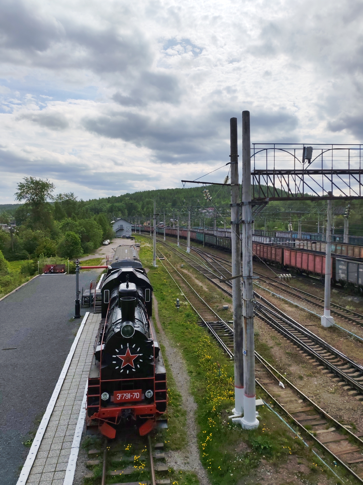 The image captures an expansive railway scene from an elevated viewpoint, likely a bridge or overpass, offering a broad perspective over multiple tracks. In the foreground on the left, a black steam locomotive with a red five-pointed star on its front and the designation "3Э791-70" in white text on a red plate is prominently featured. It is positioned on a track alongside a light-colored platform, suggesting it might be a display piece or a historical artifact at a station.
To the right of the locomotive, a complex network of railway tracks stretches into the distance, accommodating several long freight trains, some appearing to be stationary or moving slowly. The freight cars are diverse in color, including red, green, and grey. Tall concrete poles with intricate overhead catenary systems and support structures line the tracks, indicating an electrified railway. A large, lattice-like metal structure, possibly part of the overhead power system or a pedestrian bridge, is visible on the right.
The surrounding landscape is characterized by lush green vegetation, including dense forests covering hills in the background, suggesting a rural or semi-rural setting. A few distant buildings and some parked cars are visible amidst the trees on the left side, hinting at a small settlement or railway outpost. The sky is overcast with numerous grey and white clouds, but the overall lighting suggests it is daytime. No people are visible in the image. The scene conveys a sense of industry and transportation in a natural, forested environment, specifically Medvezhyegorsk, Russia.