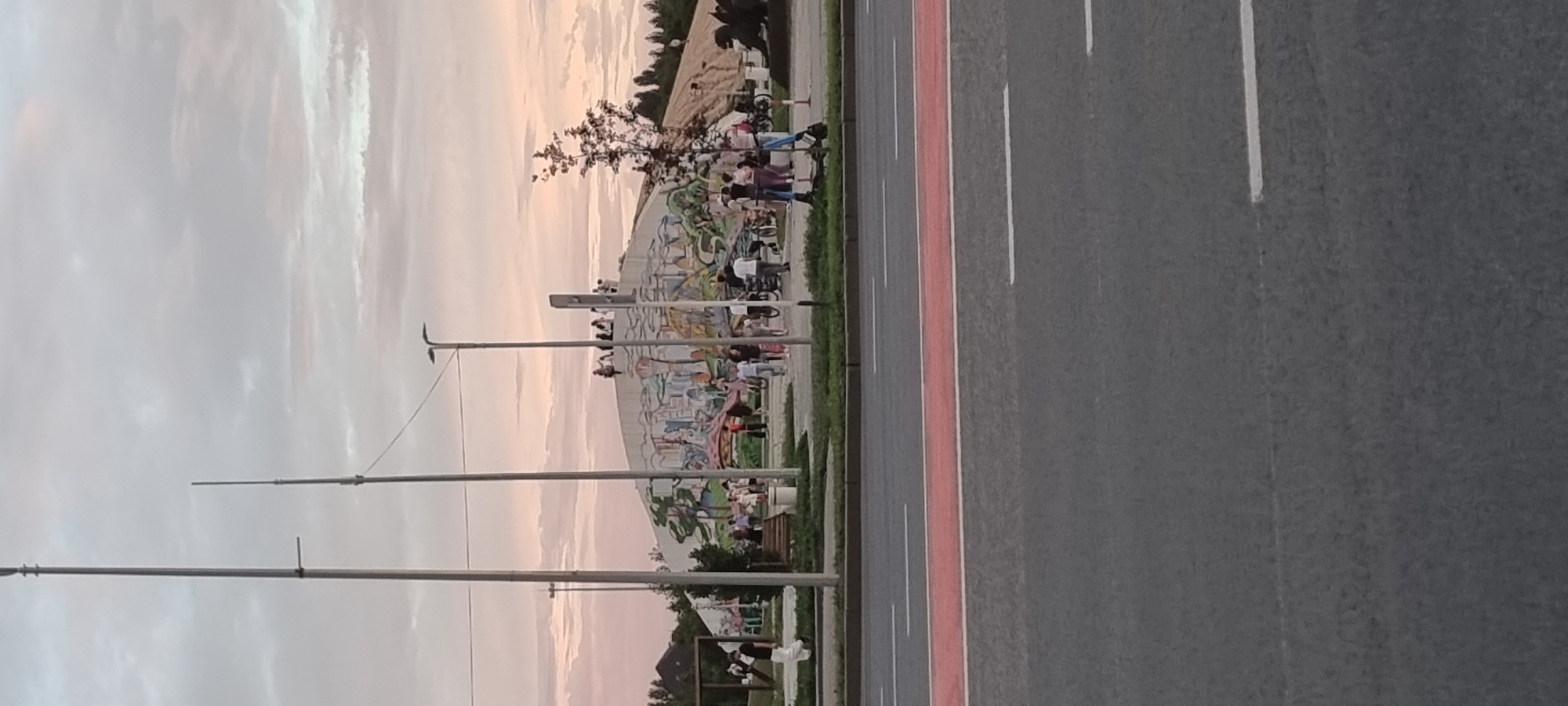 The image depicts a street scene in Aktobe, Kazakhstan, with a large, mural-covered structure in the background. The sky is overcast with muted, pastel colors. In the foreground, a grey asphalt road is visible with a red lane marking and white lane lines. Behind the road, there is a grassy area and a paved walkway where several people are gathered. Some individuals are standing, others are walking, and a few are on bicycles or scooters. The mural on the structure appears to be colorful and abstract, depicting various shapes and figures. Tall poles, likely streetlights or flagpoles, are scattered throughout the scene. A few trees are also present. The overall impression is a public space in a city during the late afternoon or early evening.