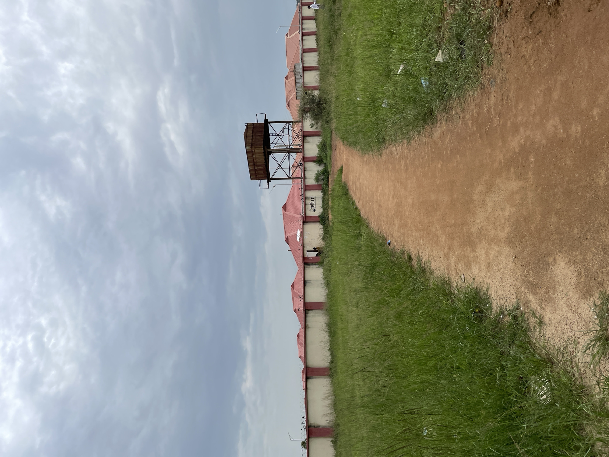 The image depicts an outdoor scene with a dirt path leading towards a compound enclosed by a wall. The compound features several buildings with red roofs, and a tall, rusted water tower stands prominently within the compound. The wall itself is light-colored with red trim and topped with barbed wire. Lush green grass grows on either side of the path, with some scattered litter visible in the grass. The sky is overcast with a cloudy, gray appearance. In the background, a street light can be seen to the left of the wall, and a sign with the text "TETFUND" is visible on the compound wall. A few individuals are present, including two standing near an entrance in the wall, and another person walking on the path within the compound on the right side. The scene suggests a residential or institutional area in Durumi, Nigeria.