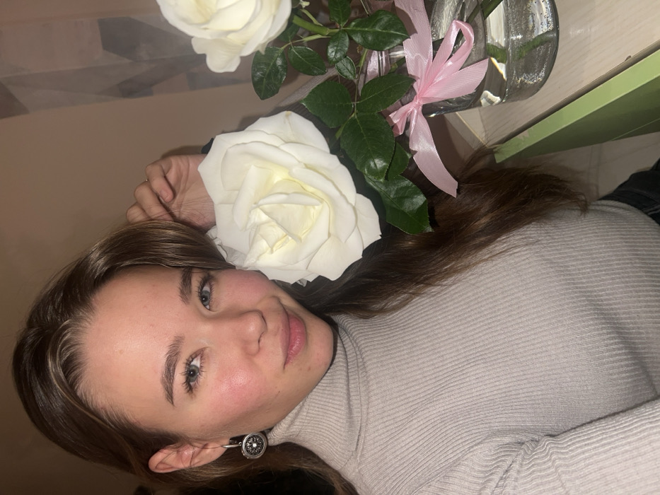 A young adult female is depicted in a close-up, indoor setting. She has light brown hair, light-colored eyes, and fair skin with visible blush on her cheeks. She wears a light beige ribbed turtleneck sweater and a circular silver earring with a dark, patterned center on her left ear. Her left hand is near her temple, touching her hair. She holds a large white rose near her face with her right hand, while looking directly at the camera with a slight smile. To her right, a clear glass vase or jar containing two white roses and green foliage is present. A small pink ribbon is tied around the vase. The background consists of a plain light beige wall on the left, transitioning to a subtle geometric pattern in shades of light gray and beige on the right. A light-colored surface is beneath the vase, with a partial view of a light green object at the lower right. The image contains no visual information to identify its specific location as Krasnoyarsk, Russia.