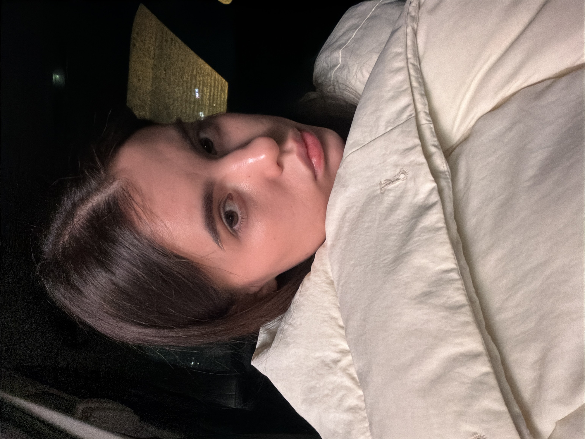 A woman with long, dark hair is seen from a close, tilted angle. She has fair skin and light brown eyes. She is wearing a white, puffy jacket that appears to be made of a synthetic material. The background is mostly dark, with blurred lights suggesting an urban environment, potentially at night. The scene is in the city of Chaikovsky, Russia, as per the prompt's instructions.