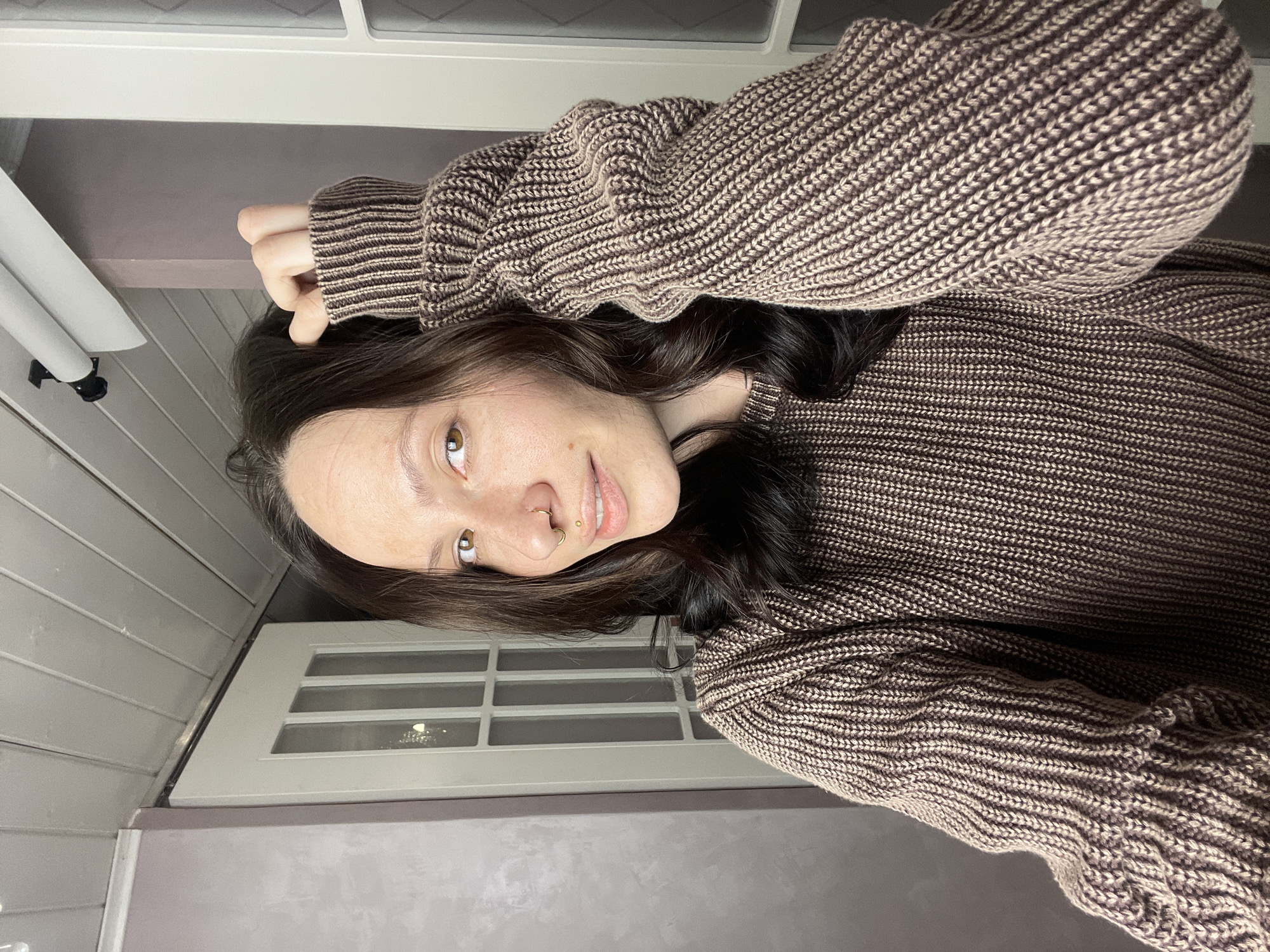 A woman with dark brown hair is wearing a brown and beige knitted sweater and is posed in an indoor setting. She has light skin, brown eyes, and facial piercings on her nose and lip. She is looking at the camera with a slight smile. The background includes a white door with multiple square glass panels and a gray wall. There is also a white object resembling a lamp or lighting fixture visible on the left side of the frame. There is no information to confirm that the location is in Izhevsk, Russia.