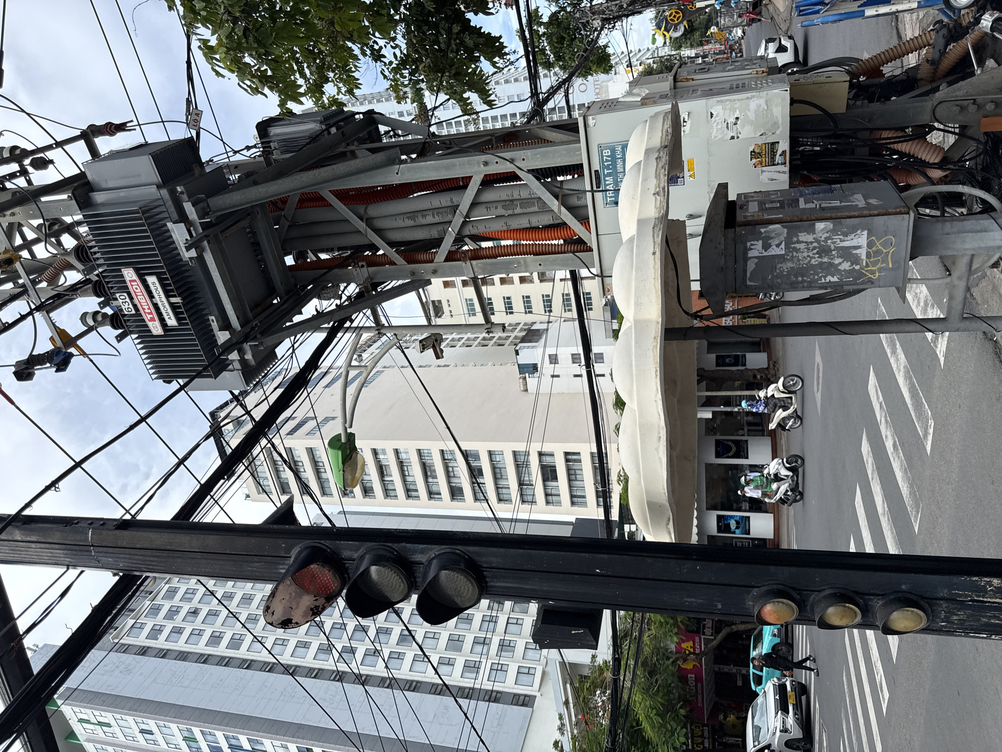 The image depicts a street scene in Nha Trang, Vietnam, focusing on a traffic light pole and a large electrical transformer. The traffic light, positioned on the left, shows a red light illuminated. Behind it, tall modern buildings rise against a cloudy sky. A prominent electrical transformer, labeled "THIBIDI" and "AMORPHOUS," is mounted on a metal utility pole on the right, surrounded by a dense network of electrical wires and conduits. The transformer appears to be a unit with a model number "630."
The street level shows a crosswalk with white stripes. A white SUV and a turquoise vehicle are visible on the road. Two individuals on scooters are moving through the intersection. A pedestrian is crossing the street. A white, dome-shaped structure, possibly a sunshade or part of an advertisement, is situated above the electrical equipment. A grey utility box, covered in stickers and graffiti, is mounted on the pole below the transformer. The overall environment suggests a busy urban intersection with significant electrical infrastructure.