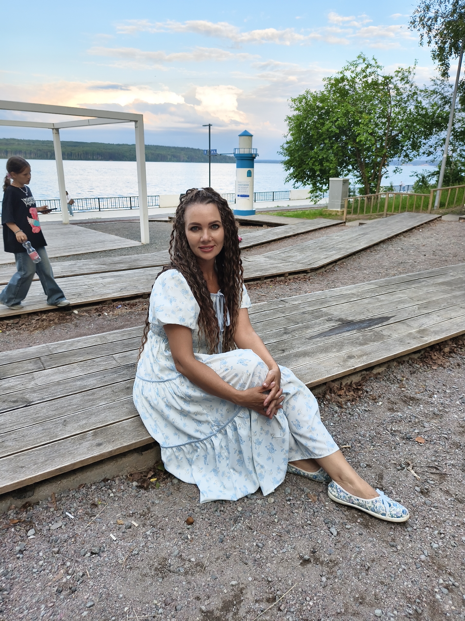 In Chaikovsky, Russia, a woman with long, curly brown hair sits on wooden benches outdoors. She wears a light blue dress with a floral pattern and matching shoes. To her left, a young girl in a black t-shirt and jeans walks away from the camera, holding a bottle. In the background, a body of water stretches towards a tree-lined horizon, with a small lighthouse visible on the shore. The scene is illuminated by natural light under a cloudy sky.