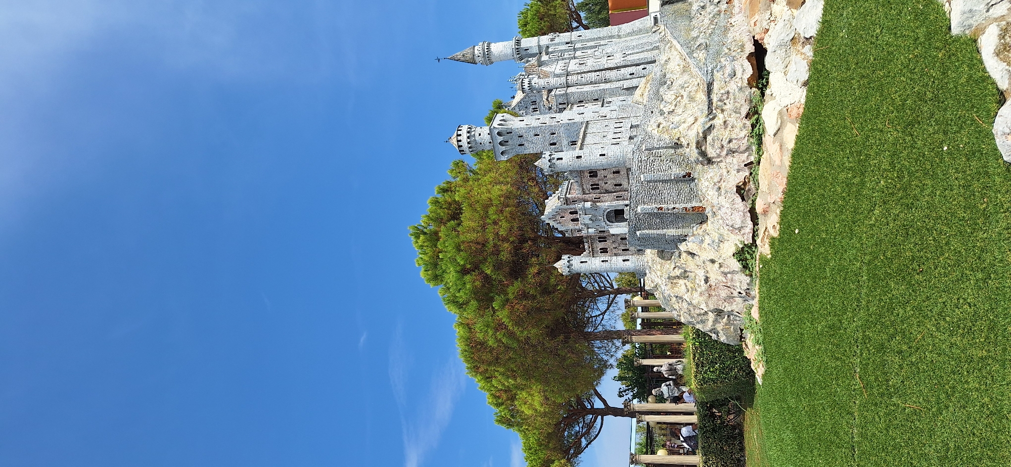 This is a photograph of a detailed miniature model of a medieval-style castle, situated on a rocky outcrop. The castle features multiple towers, some with conical roofs, and intricate stonework. In the background, a dense, green pine tree provides a natural backdrop, with a clear blue sky above. To the left of the castle, there is a garden area with manicured hedges, classical-style columns, and statues, suggesting a park or decorative garden setting. A few people are visible in the background near the garden area, appearing to be visitors. The foreground consists of a well-maintained green lawn. This miniature castle exhibit is located within the Italia in Miniatura theme park in Rimini, Italy.