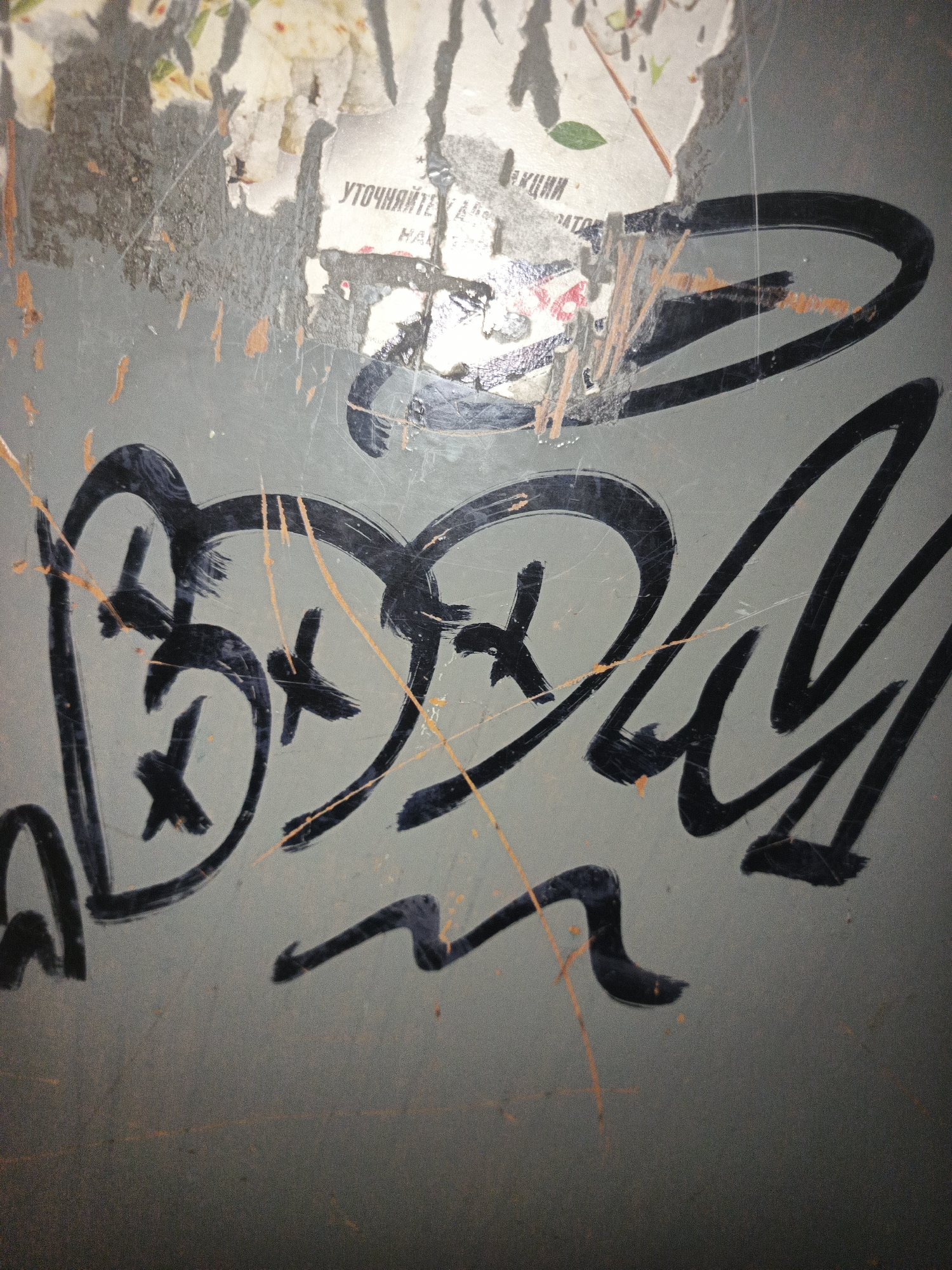 The image shows graffiti spray-painted on a grey, weathered surface, possibly a wall or a metal object. The graffiti is in black and appears to be a stylized word or symbol, with rounded shapes and sharp angles. There are also orange and yellow marks on the surface, which might be part of the graffiti or damage. In the background, there are torn pieces of paper with text and images, suggesting the location may be an urban environment with posters or advertisements. One piece of paper has the Cyrillic text "УТОЧНЯЙТЕ". The overall impression is one of urban decay and street art. The specific location in Smolensk, Russia, is not identifiable from the visual information provided.