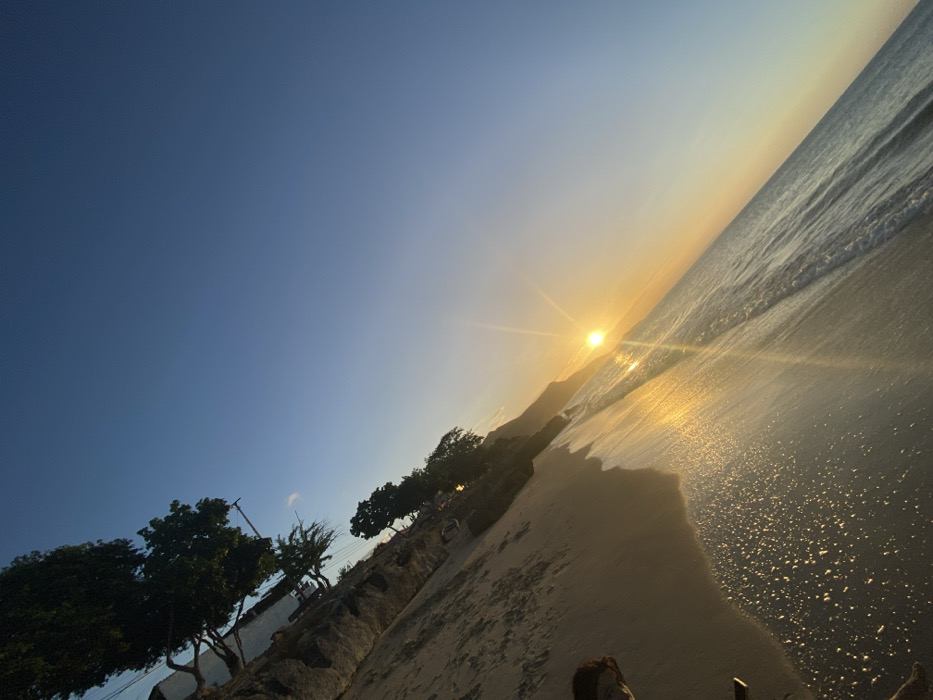 A beach scene unfolds at sunset. The sun casts a golden glow on the sand and water, reflecting intensely. Gentle waves lap the shore. Along the beach's edge, trees and rocky formations create a barrier. The sky transitions from a deep blue to a light orange near the horizon. A sense of tranquility pervades the scene.