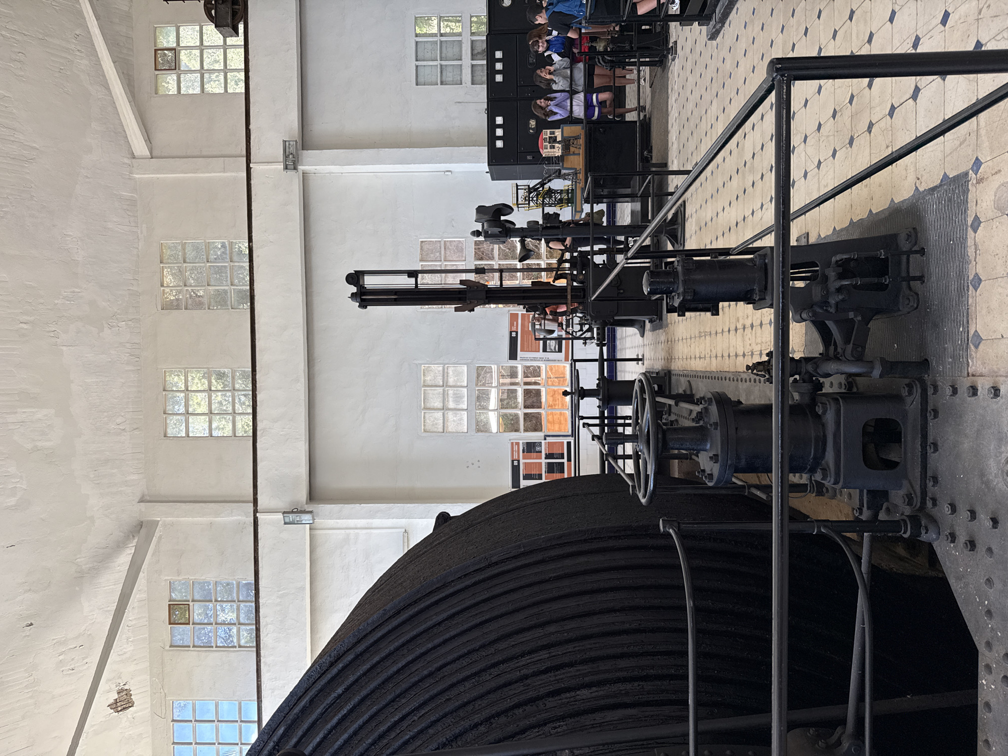 The image depicts the interior of a museum or industrial heritage site in Cardona, Spain, likely the Salt Mountain Cultural Park museum. In the foreground, a large, dark, ribbed cylinder, possibly a winch or winding drum, dominates the left side of the frame. Adjacent to it is a complex piece of industrial machinery, featuring gears, pipes, and a large handwheel. A black metal railing separates the viewer from the machinery. In the background, several people, including a group of young individuals, are observing exhibits. The space has high ceilings, white walls, and numerous windows with a grid pattern of opaque glass panes. The floor is tiled with a light color and dark blue diamond-shaped patterns. The overall impression is of a well-preserved industrial artifact within an educational setting.