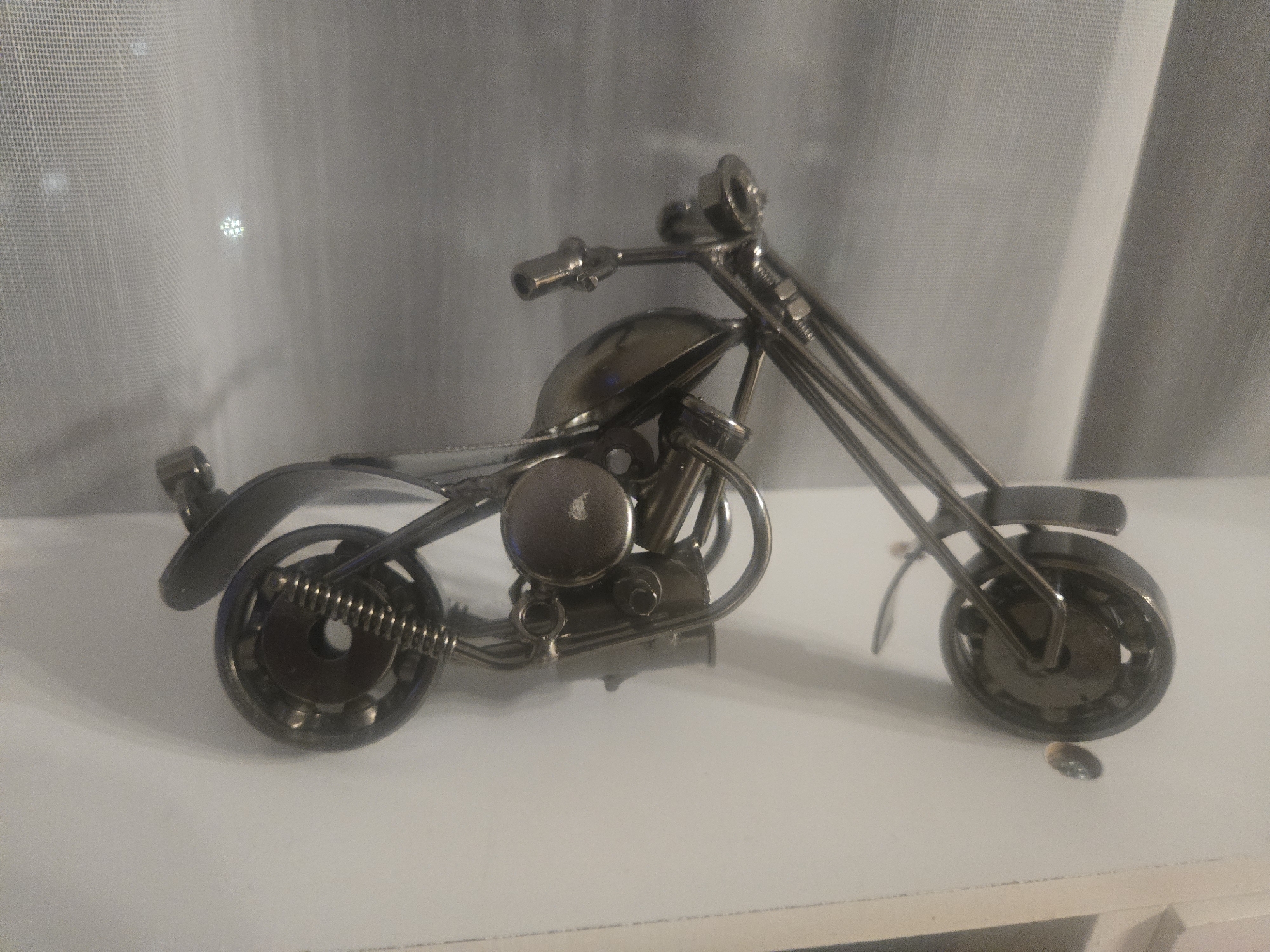 A detailed, silver-colored metal motorcycle figurine sits on a white surface. The background features a soft-focus white curtain. The location is Unknown, Unknown.