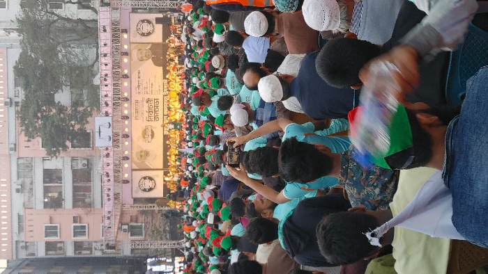An outdoor political rally is depicted in an urban area of Dhaka, Bangladesh, during daylight hours. A large crowd, primarily composed of adult males, is assembled, facing a substantial temporary stage structure at the far end of the scene.
The stage features a large banner displaying photographic portraits of Bangabandhu Sheikh Mujibur Rahman on the left and Sheikh Hasina on the right. Bengali text on the banner prominently reads "নির্বাচনী জনসভা" (Election Rally), with other visible text including "বাংলাদেশ আওয়ামী লীগ" (Bangladesh Awami League) and "শেখ হাসিনা" (Sheikh Hasina). The stage is equipped with extensive lighting rigs and large speaker arrays. Several individuals in yellow-orange attire are visible on the stage platform.
In the background, behind the stage, a multi-story, light-colored urban building with numerous windows and visible air conditioning units is present, partially obscured by bare trees.
The foreground and mid-ground are densely populated by attendees. Many individuals wear light blue t-shirts and caps featuring green and red colors, indicative of party affiliation. Some wear white kufi caps. One male individual in the mid-foreground, wearing a light blue t-shirt, holds a black smartphone horizontally above his head, directed towards the stage, capturing the event. Another male in the immediate foreground, wearing a multi-colored cap, holds a clear plastic water bottle, exhibiting motion blur. An individual wearing an orange high-visibility vest is visible in the crowd on the right. A white fabric object, possibly a small flag, is visible in the lower-left.