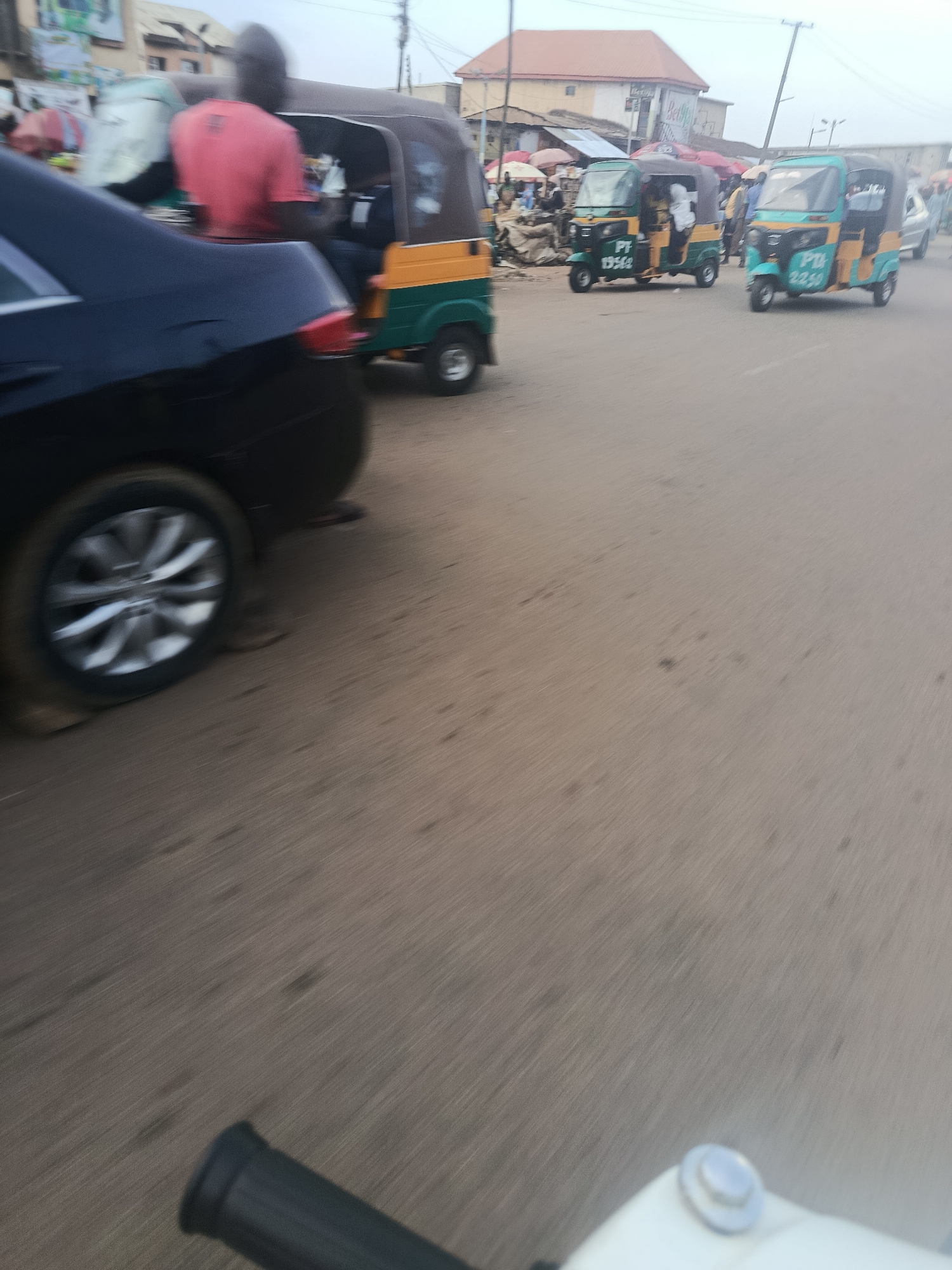 This is a motion-blurred, eye-level, outdoor photograph taken from the perspective of a motorcycle or similar vehicle, looking towards a street scene in Rafin Par, Nigeria. The foreground is dominated by the handlebar and part of the seat of the vehicle the photographer is on, with a white object and a silver fastener visible on the seat.
The midground shows a black car with a silver wheel in motion, its movement emphasized by the blur. Behind the car, several auto-rickshaws, known locally as Keke Napeps, are also in motion. Two of these are turquoise and yellow, with numbers "PT 19562" and "PT 2259" visible on their sides. Another auto-rickshaw, predominantly green and yellow, is partially obscured by the black car. A person wearing a pink shirt is visible in the driver's seat of the green and yellow auto-rickshaw.
The background depicts a busy street with various structures, including buildings with red roofs, market stalls with colorful umbrellas, and overhead wires. Several people are visible in the background, blurred due to motion and distance. The overall scene suggests a bustling urban environment in Nigeria, likely a market area, with active transportation and commerce.