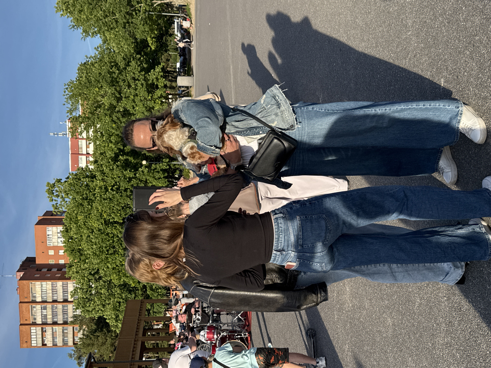 A group of people are gathered outdoors, with a woman in the foreground holding a small dog. She is wearing a denim jacket and wide-leg jeans. To her left, another woman in a black long-sleeve shirt and blue jeans stands with her arm around the woman holding the dog. A third person in a black leather jacket is partially visible on the far left. In the background, there are trees, buildings, and what appears to be a drum set for a performance. The sky is blue and clear, suggesting a sunny day. This scene is located in Tallinn, Estonia, possibly at an outdoor event or gathering.