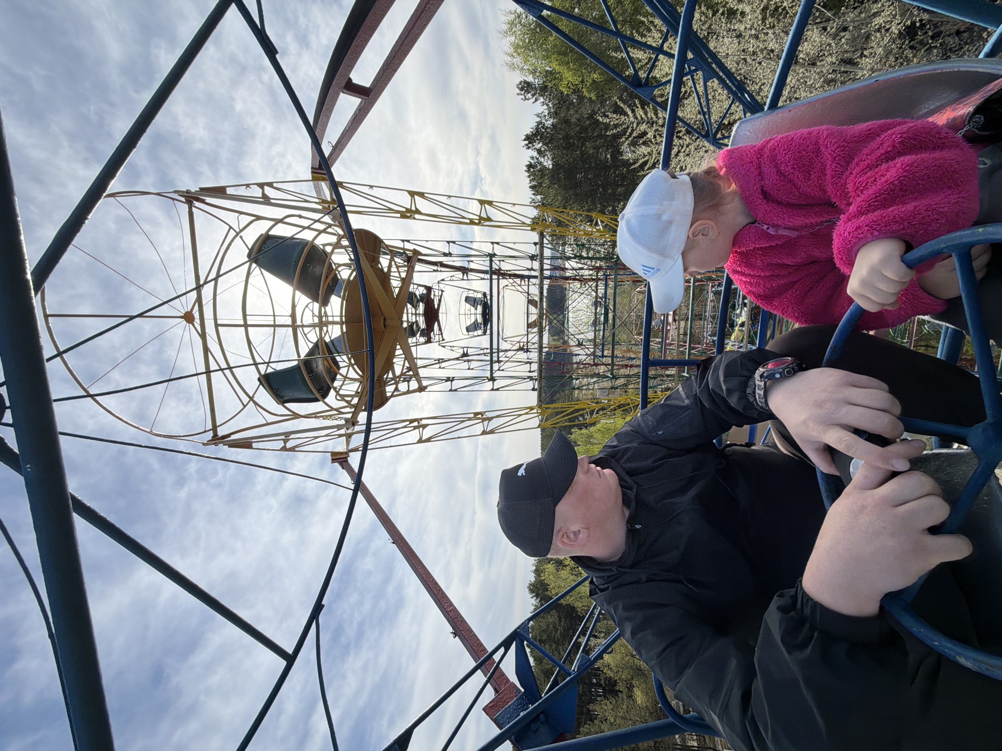 The image depicts a low-angle view of a Ferris wheel with a man and a child riding in one of its carriages. The location is the city of Chaikovsky, Russia.
The Ferris wheel's structure is made of metal, with a combination of yellow, red, and blue supports. The carriages are yellow and green.
A man is sitting in the carriage, wearing a black jacket and a black baseball cap. He is holding onto the railing.
Next to him is a child, who is wearing a pink fleece jacket and a white baseball cap. The child is also holding onto the railing of the carriage.
The background shows a sky with scattered clouds and greenery, including trees. The image suggests the Ferris wheel is located in an outdoor park or amusement area.