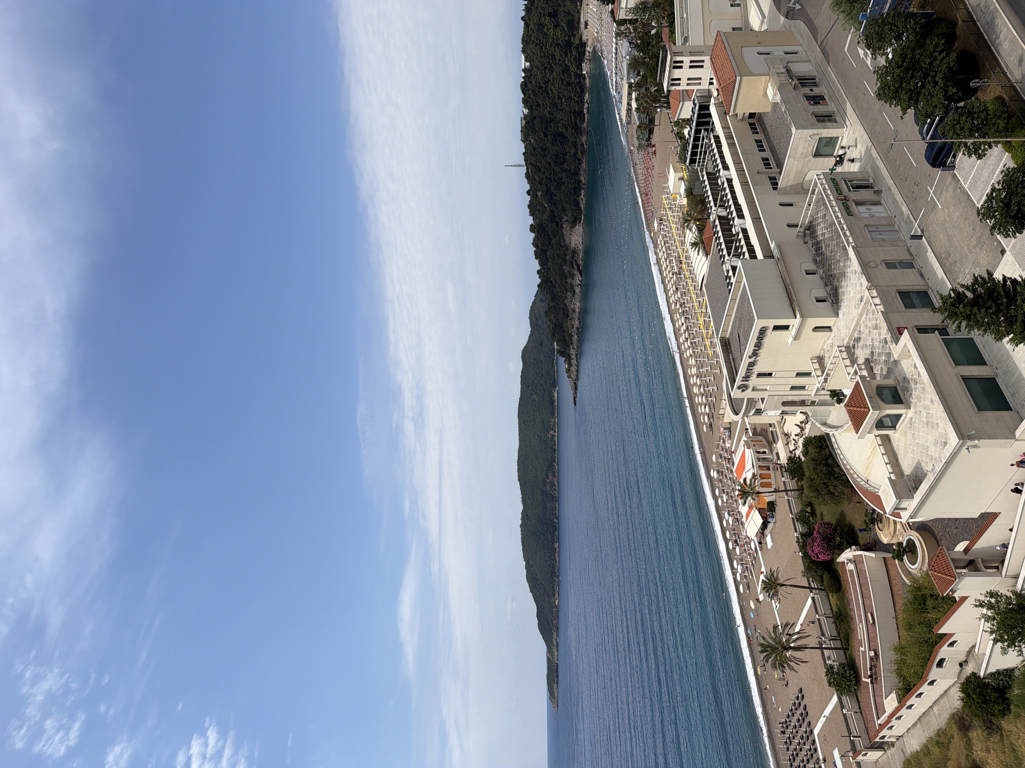 This image, set in Boreti, Montenegro, captures a vibrant coastal resort scene under bright daylight. The primary view is a long stretch of beach and the calm, deep blue sea, bordered by buildings on one side and a lush, forested hill on the other.
The foreground features a wide, sandy beach populated with numerous rows of white sun loungers, many topped with yellow umbrellas. Some darker sun loungers are also visible further down the beach. A few people can be seen enjoying the beach and water, indicating a leisure or vacation atmosphere. The sea appears tranquil, with visible buoys or markers delineating areas in the water.
On the right side of the image, a series of modern, multi-story buildings line the coast, likely comprising hotels or resorts. These structures are predominantly light-colored with reddish-brown tiled roofs and feature balconies and terraced areas. Prominently visible on one building is the text "HOTEL SPLENDID". Further along, another building displays a partially visible sign that appears to read "ADRIATIC BEAUTY RESORT". A paved road runs parallel to these buildings, where a few cars are parked. Palm trees and other green foliage are scattered throughout the resort area, adding to the picturesque setting.
To the left, across the expanse of blue water, a large, dark green forested hill rises steeply from the sea, extending into the distance. A tall, slender structure, possibly a communication mast or tower, is faintly visible atop this distant hill. The sky overhead is a clear, bright blue with scattered white, wispy clouds, suggesting excellent weather conditions.
The overall scene depicts a sunny day at a popular coastal destination, bustling with activity but maintaining a sense of relaxation.