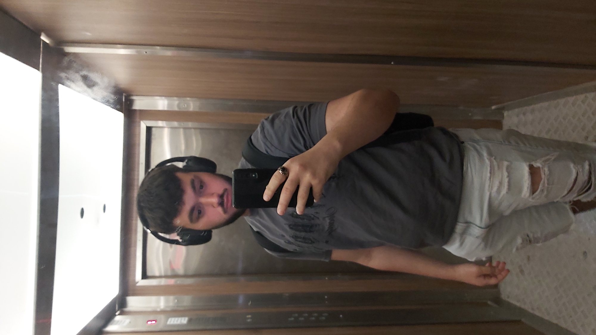 A person is taking a selfie in what appears to be an elevator. The person is wearing headphones, a gray t-shirt, and ripped light-colored shorts. They are holding a phone in their right hand, which is capturing the image. The elevator walls are metallic with wood trim. The floor is tiled. There is not enough information to ascertain the location in Sofia, Bulgaria.