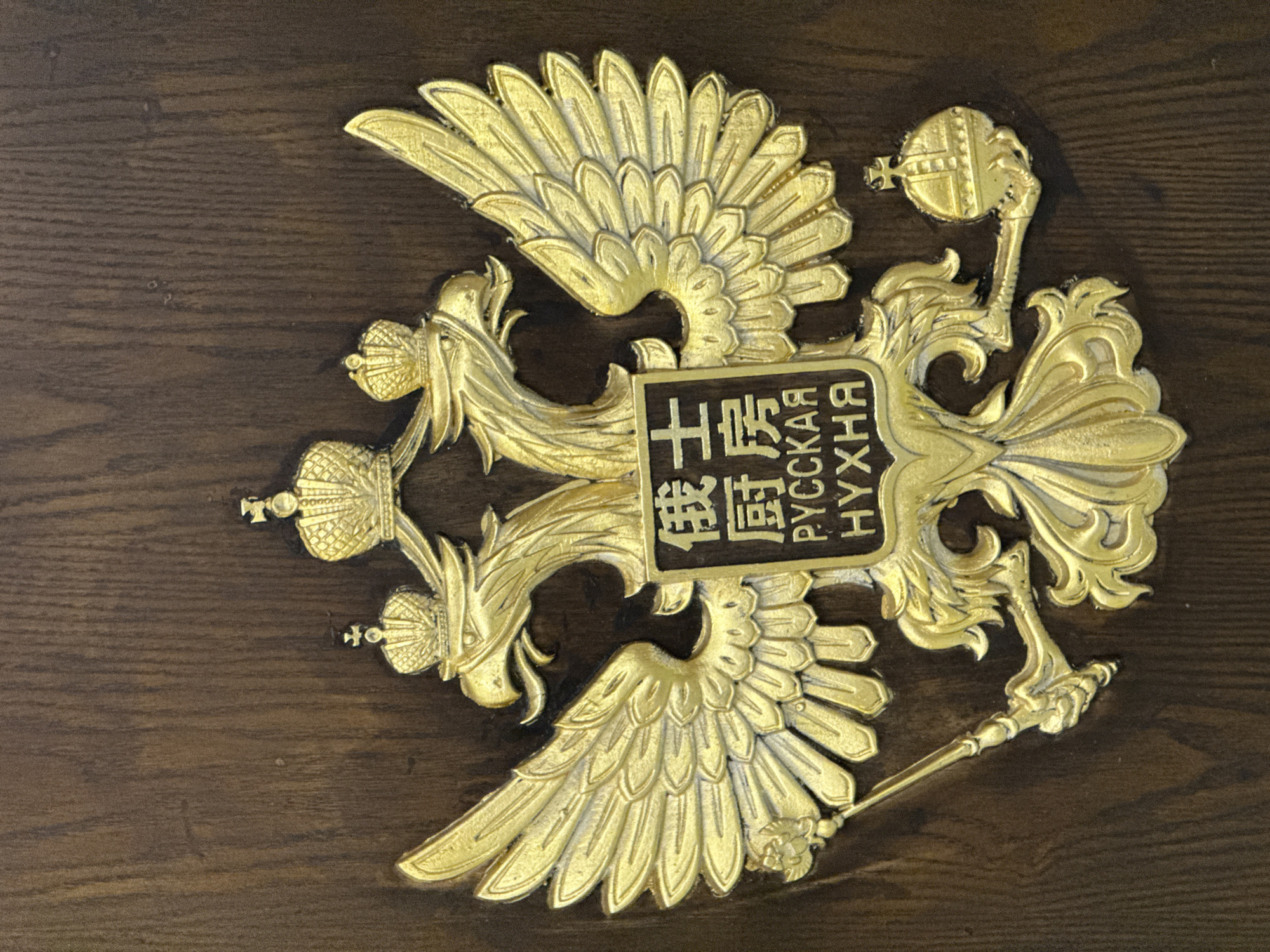 A close-up shot shows a gold-colored, ornate emblem affixed to a dark, wood-grained surface. The emblem features a double-headed eagle design, a common symbol used in various cultures and nations. It includes crowns atop each head, a scepter in one claw, and an orb in the other. A rectangular plaque is centered on the eagle's chest, with lettering in both Chinese and Cyrillic script. The words "РУССКАЯ КУХНЯ" (Russian Kitchen) are written in Cyrillic. The location of Tianhe District in China is referenced in this image via the Chinese lettering on the rectangular plaque.