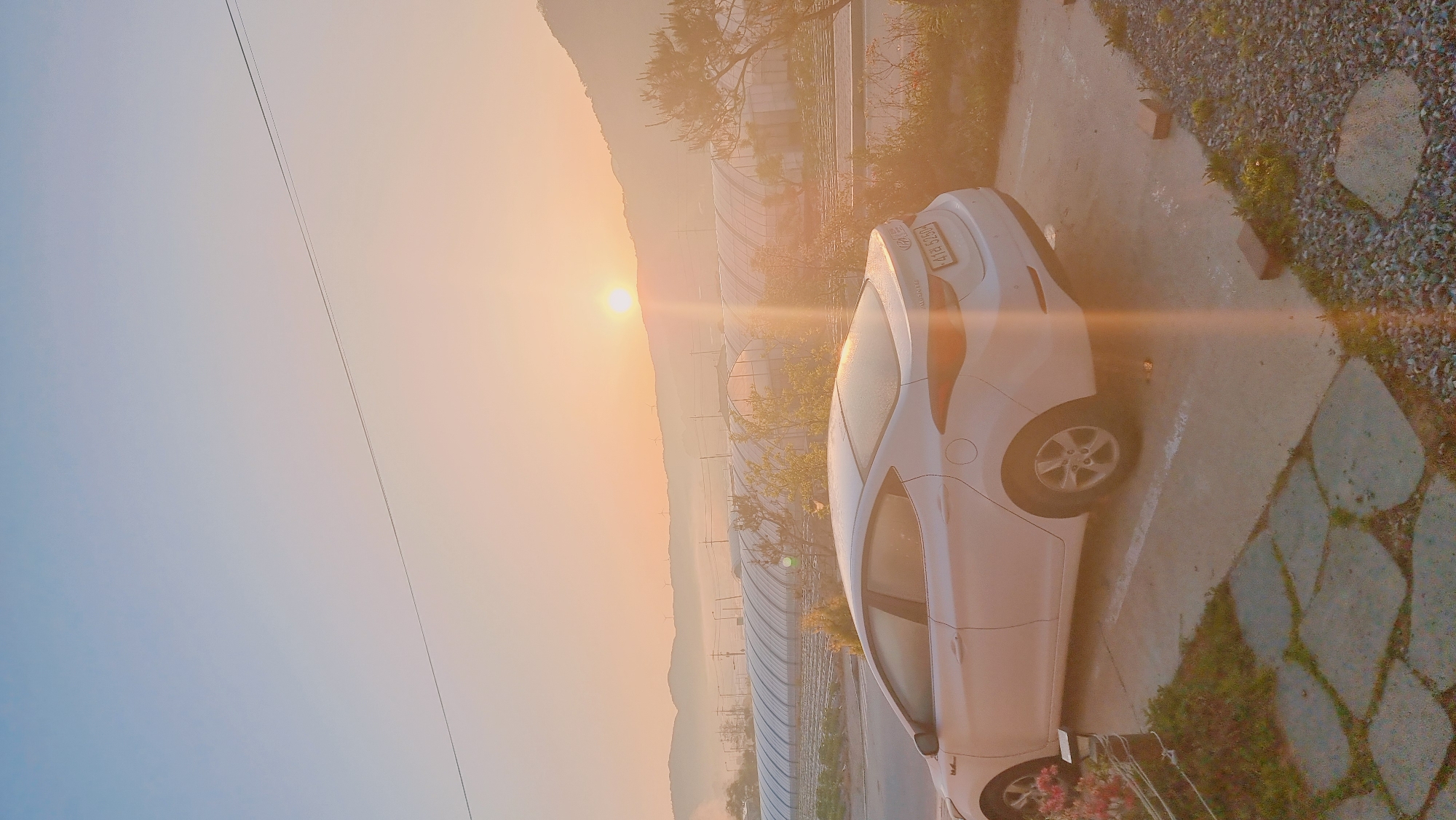 The image shows a white Hyundai Elantra parked on a concrete driveway adjacent to a stone and gravel walkway. The driveway is next to a road lined with agricultural greenhouses. In the background, hills and the sun are visible through a hazy atmosphere. The car's license plate reads "4178 5250". The location is Dunnae-myeon, South Korea. The sun is low on the horizon.