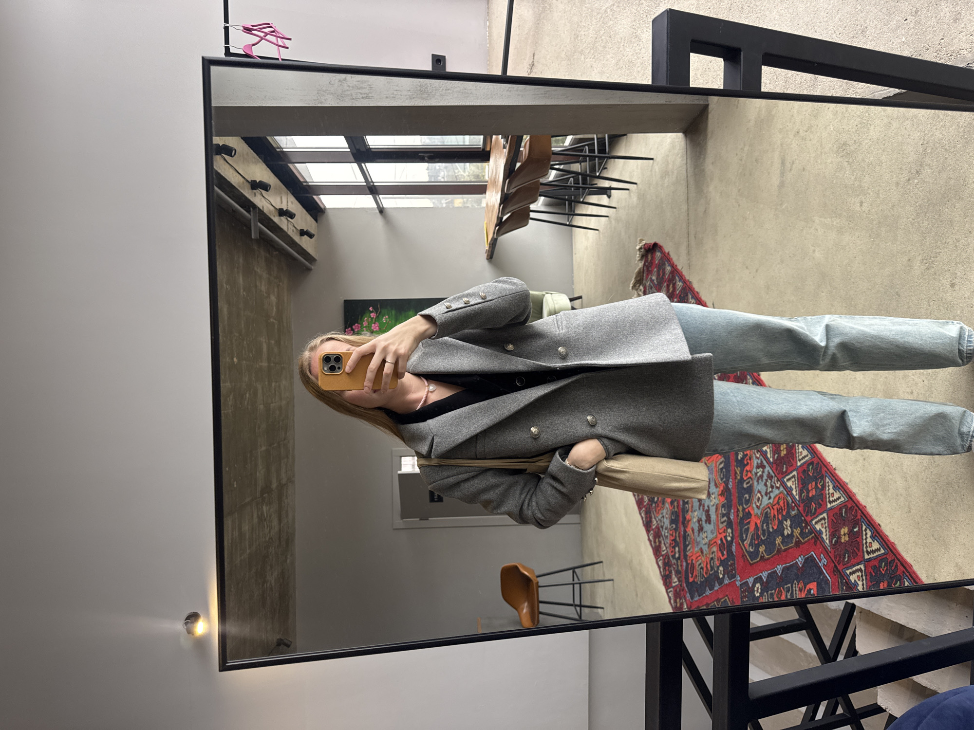 A woman is taking a selfie in a mirror. The mirror reflects a modern interior space with a high ceiling, exposed concrete beams, and large windows. Reflected in the mirror are chairs and a table, a patterned rug, and a painting with floral motifs. The woman is wearing a gray blazer, a dark top, and light-wash jeans. She is holding a phone with a yellow case. A pink hanger is visible on top of the mirror. The background suggests an urban loft or studio space. There is no indication this image was taken in Sochi, Russia.