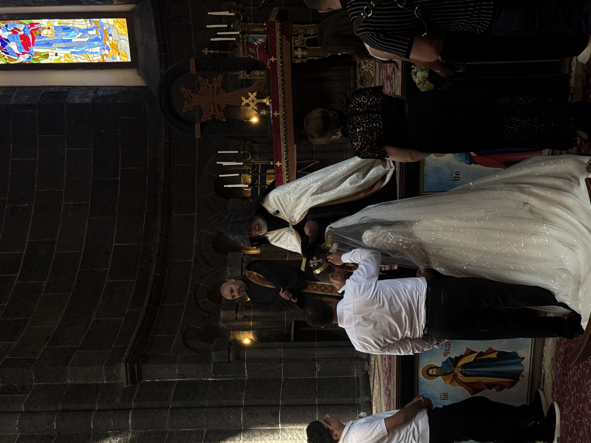 This image depicts a wedding ceremony taking place inside a dimly lit stone church, likely in Gyumri, Armenia, given the religious context and architectural style often found there. A bride in a sparkling white gown and veil stands with her groom, who is wearing a white shirt. A priest, distinguished by his ornate white and gold vestments and black head covering, officiates the ceremony, holding a book. Another clergyman in a black robe stands behind them. Several other individuals are present, observing the ceremony. In the background, an altar with candles and a crucifix is visible, along with a stained-glass window depicting a religious figure. The environment is characterized by rough stone walls and a patterned carpet.