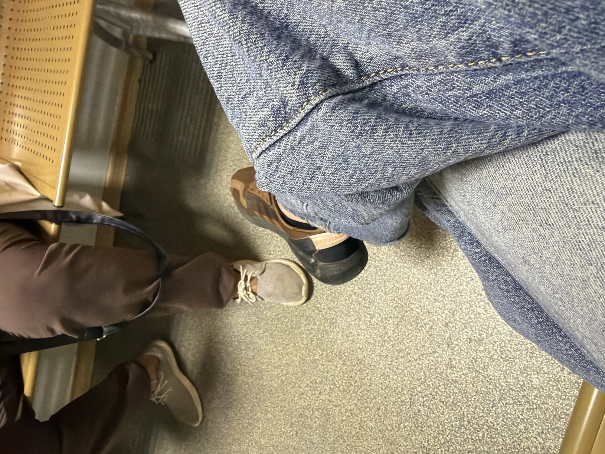 This eye-level shot captures an indoor scene, likely a public waiting area, in Chaikovsky, Russia. The perspective looks down towards the floor, showing the lower halves of two individuals seated.
On the right, one person is wearing light blue denim jeans, notable for their visible texture and yellow stitching. They are wearing dark brown shoes with lighter brown uppers and black soles. A white label with black text, partially decipherable as "MARQUE ALL", is visible on the heel of one shoe. To the left, another individual wears brown trousers and light grey lace-up sneakers with white laces.
Part of a light brown or tan chair with a perforated backrest is visible on the far left, suggesting public seating. A black bag is partially obscured beneath the brown-trousered leg. The floor is a light, speckled material, possibly short-pile carpet or linoleum, common in public spaces. The individuals appear to be stationary, indicating a moment of waiting or rest. No specific time of day or weather can be determined from this indoor view.