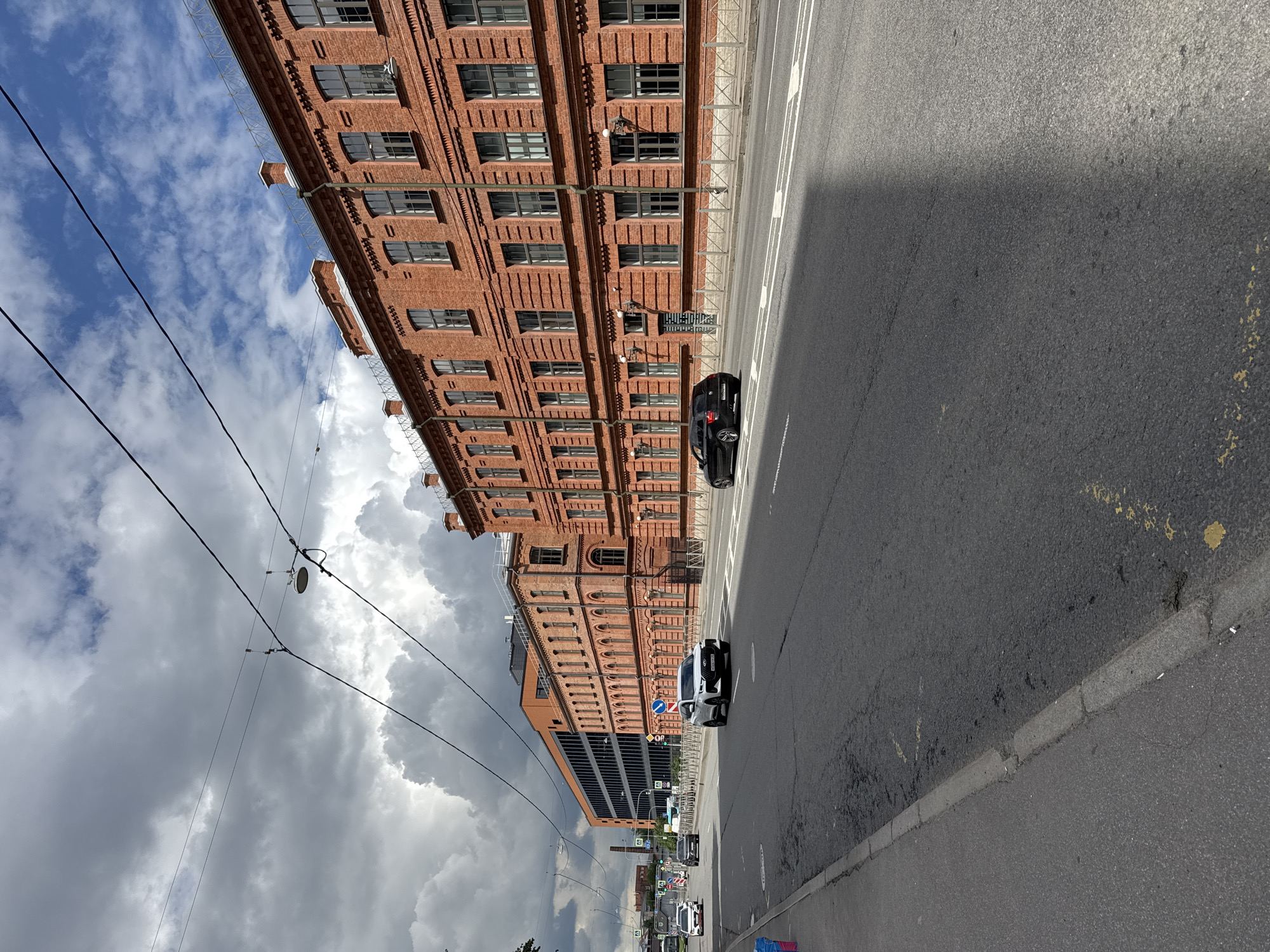 The image captures a street scene in Saint Petersburg, Russia, on a cloudy day. The foreground features a two-lane asphalt road with white lane markings and a gray concrete curb on the left. The road surface shows signs of wear with some patches and yellow markings. On the right side of the road, a row of red brick buildings with numerous windows lines the street. To the left of these buildings is a modern, darker building with a dark facade and windows. Cars are visible on the road, including a white SUV in the right lane and a dark sedan in the left lane. Overhead, power lines and a street lamp are strung across the frame against a sky filled with a mix of dark, ominous clouds and patches of blue with scattered white clouds. The overall impression is an urban environment characterized by historical and modern architecture under a dynamic sky.