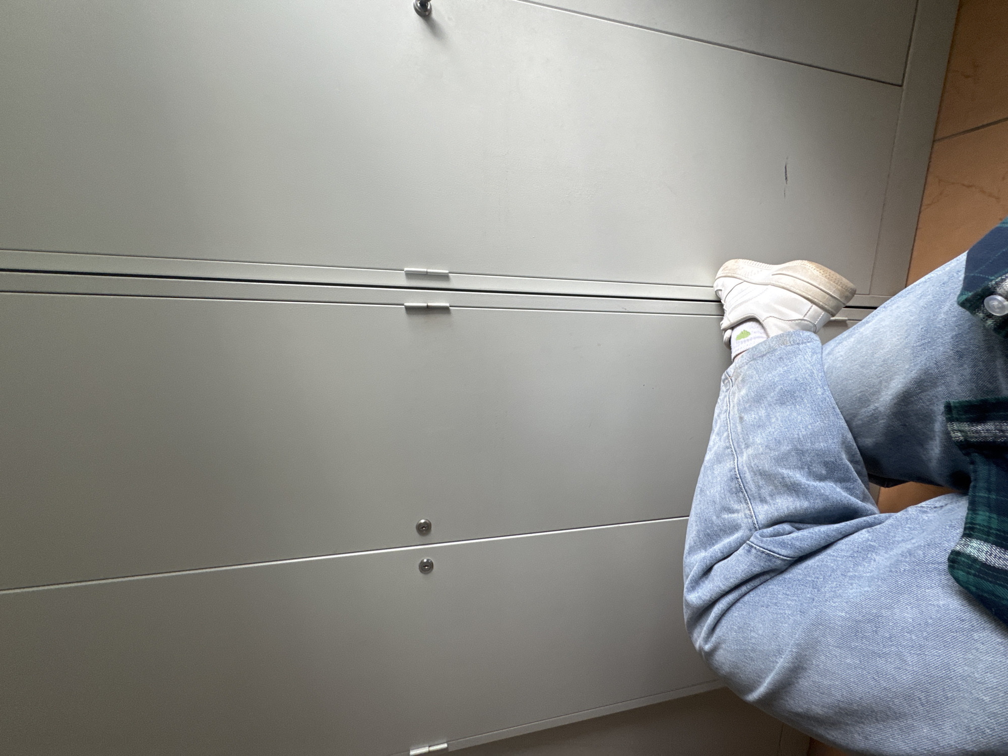 A person is sitting with their legs crossed in front of a light gray metal filing cabinet. The person is wearing light blue jeans and white sneakers with green accents. A blue and green plaid shirt is visible on their arm. The filing cabinet has multiple drawers, indicated by horizontal lines and visible drawer pulls. Two small, round indentations, possibly locks or handles, are visible on the front of the cabinet. The scene appears to be indoors, likely an office or storage area, given the filing cabinet. The lighting suggests it could be daytime, but there are no strong indicators of the time of day or weather. No text is visible in the image. The setting is identified as Perm, Russia.