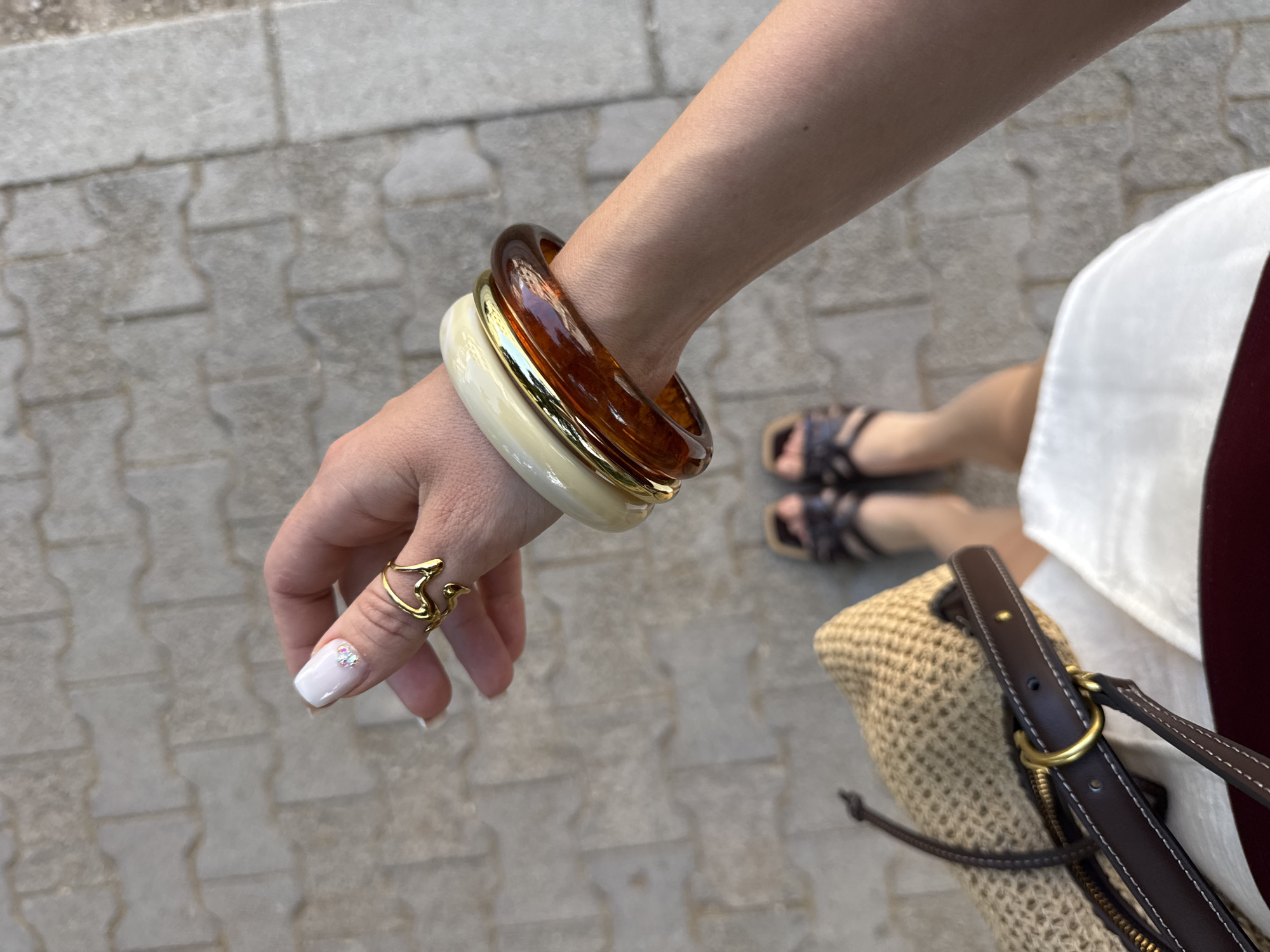 The image is a close-up, overhead shot of a woman's hand and arm, adorned with jewelry, as she stands on a cobblestone walkway. Her fingernails are painted white with small iridescent rhinestones on the ring finger's nail. She wears a unique gold ring on her right index finger, shaped like a fluid, abstract heart or drop. Around her wrist are three chunky bangles: one appears to be a translucent cream color, another a metallic gold, and the outermost is a rich, dark amber tortoiseshell pattern. Her hand is positioned as if reaching towards a straw tote bag with dark brown leather straps and gold hardware, which is visible in the lower left of the frame. Below her arm, her legs are visible, clad in dark brown sandals with straps. The background is a neutral grey and tan cobblestone pavement, suggesting an outdoor urban environment. The lighting indicates it is daytime, with diffused sunlight. The scene is set in the city of Budva, Montenegro, a coastal town known for its historic old town and beaches.