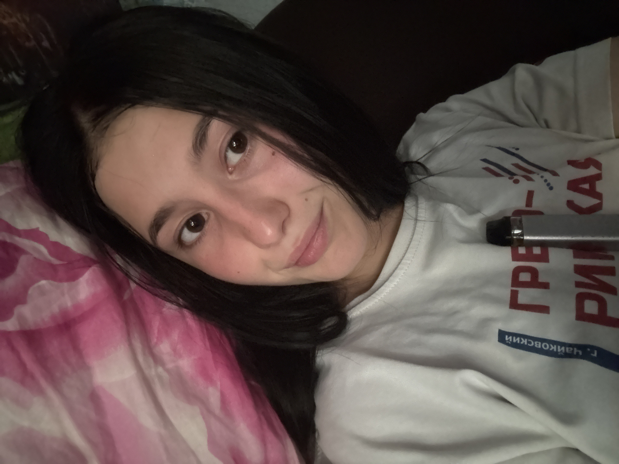 The image shows a young woman with long, dark hair lying down, possibly in bed, as evidenced by the pink and white patterned fabric she is lying on. She is looking towards the camera with a slight smile. She is wearing a white t-shirt with red and blue text printed on it. Some of the text is visible and appears to be in Cyrillic script. The overall lighting suggests it could be indoors or evening. There are no other people or significant objects in the frame besides the woman and her clothing. The setting seems personal and casual. The t-shirt has text that reads "ГРЕ" in red, followed by some blue stripes and dots, and then "РИ" and "КАS" in red. Below this, there is a blue rectangle with white text that appears to say "г. ЧАЙКОВСКИЙ", which translates to "Chaykovsky city" in Russian.