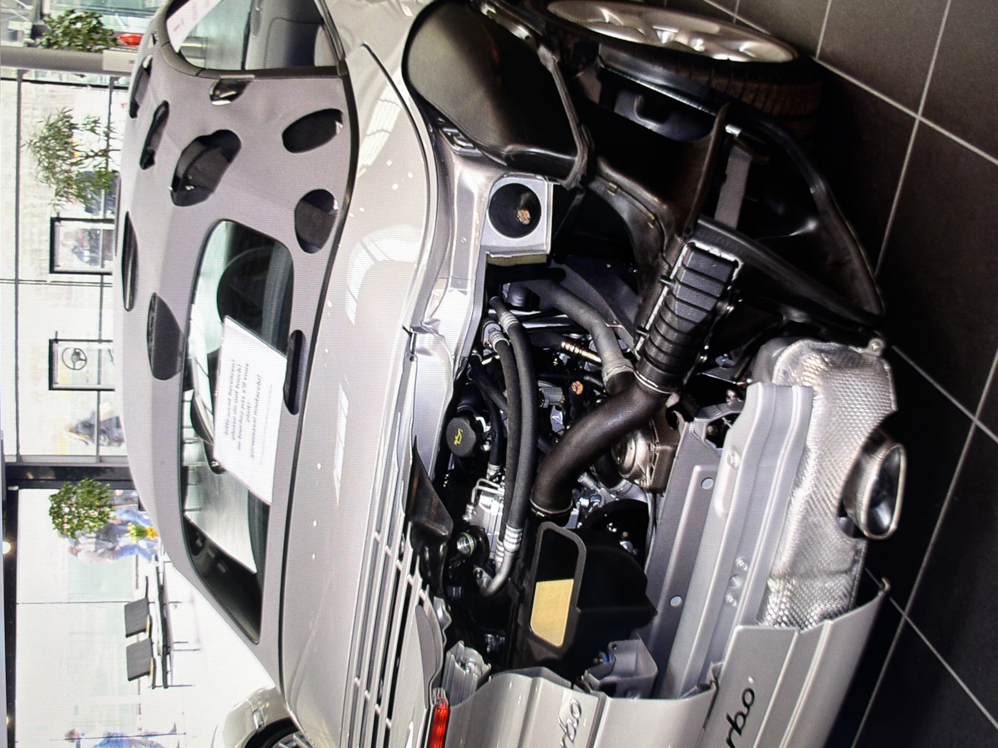 This is a close-up, eye-level, medium shot of the rear of a silver Porsche 911 convertible, displayed indoors on a dark tiled floor. The car's engine bay is exposed, revealing intricate mechanical components, including the engine, exhaust system, and air intake. The soft top of the convertible is partially down, revealing a patterned grey fabric with circular cutouts. A white sign with text is visible through the windshield. The background is blurred, showing a large window with a view of trees and people walking, suggesting a showroom or exhibition space. The image does not provide any information about the city of Kyiv, Ukraine.