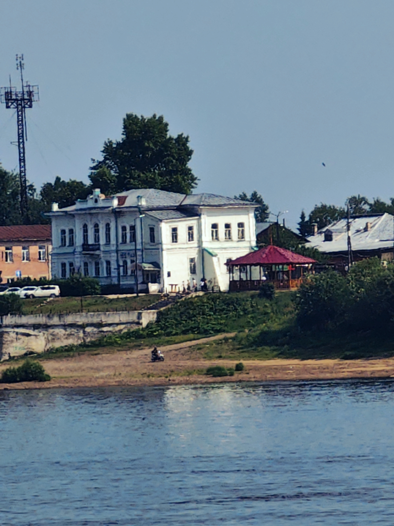 The image shows a waterfront view of Yeniseysk, Russia, with several buildings and natural elements. In the background, a tall communications tower stands against a clear blue sky. A large, light-colored, multi-story building with many windows and ornate architectural details is prominently featured. To its left, a smaller, reddish-brown building is visible. In front of the buildings, a red-roofed gazebo sits on a grassy slope. A body of water, likely a river, occupies the foreground, with reflections of the sky and surrounding scenery on its surface. The riverbank is lined with sand and vegetation. On the bank, a person is riding a motorcycle. Several white cars are parked near the buildings, suggesting a public area or road. The overall scene depicts a tranquil day in a riverside town.