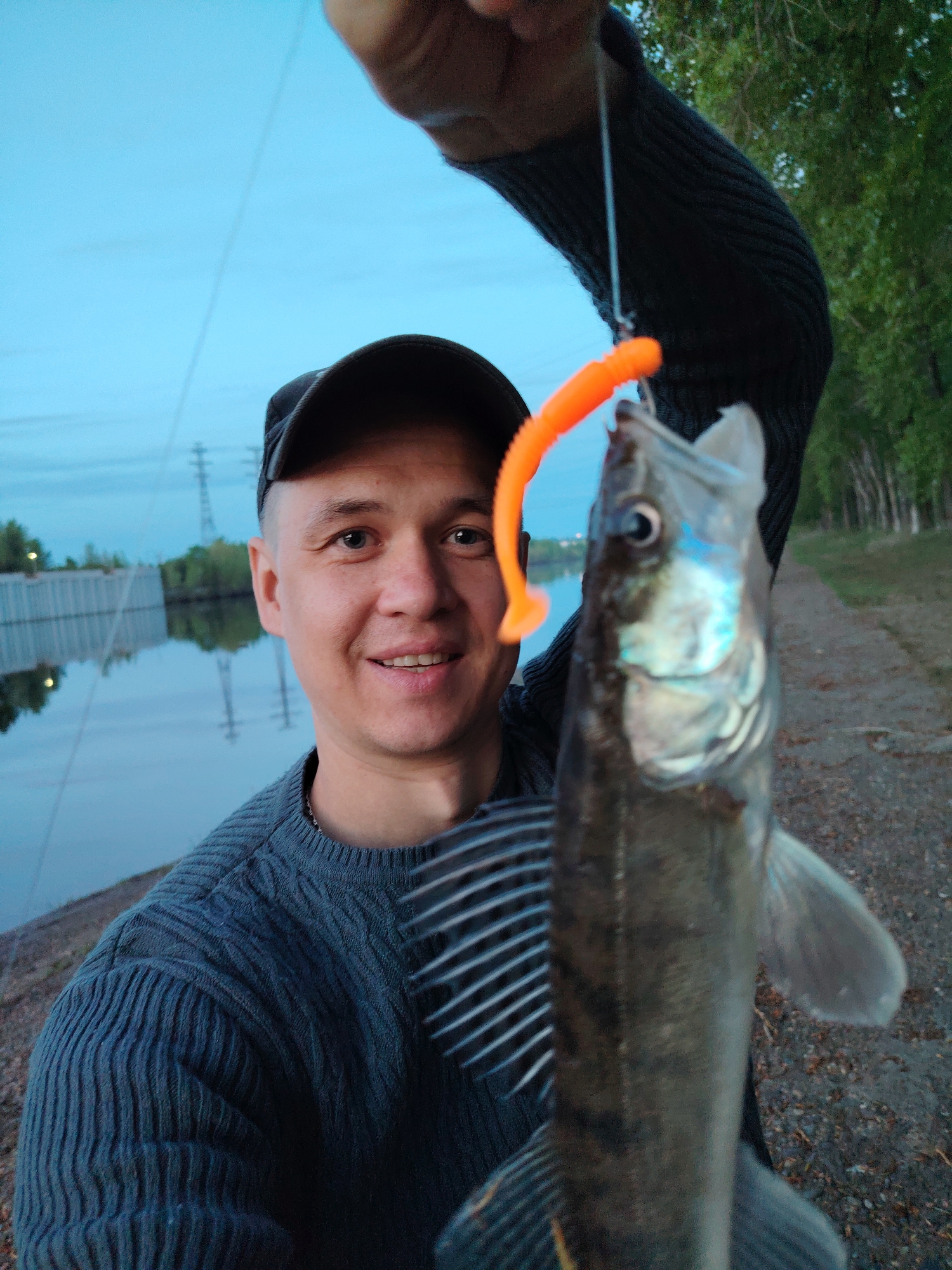 A man in a navy blue sweater and a black baseball cap is holding up a zander fish he has caught while fishing on the Kama River in Chaikovsky, Russia. The fish is hanging from fishing line, and it has an orange lure in its mouth. The river, power lines, and a river wall are visible in the background on the left. A tree line and path are in the background on the right. The man is smiling and looking at the camera.