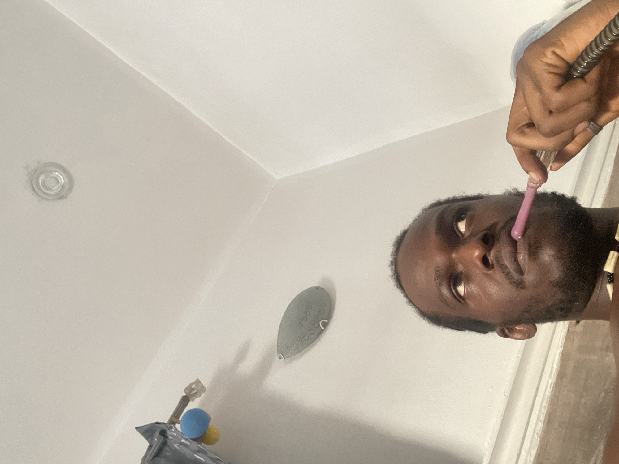 A low-angle shot depicts a man with dark hair and beard. He holds a pipe with a pink mouthpiece to his lips. His hand is visible, holding the pipe and wearing a ring. The background includes a white wall with a light fixture, a gray object with a pattern, and possibly an object with a blue ball. The location is Kuchingoro, Nigeria.