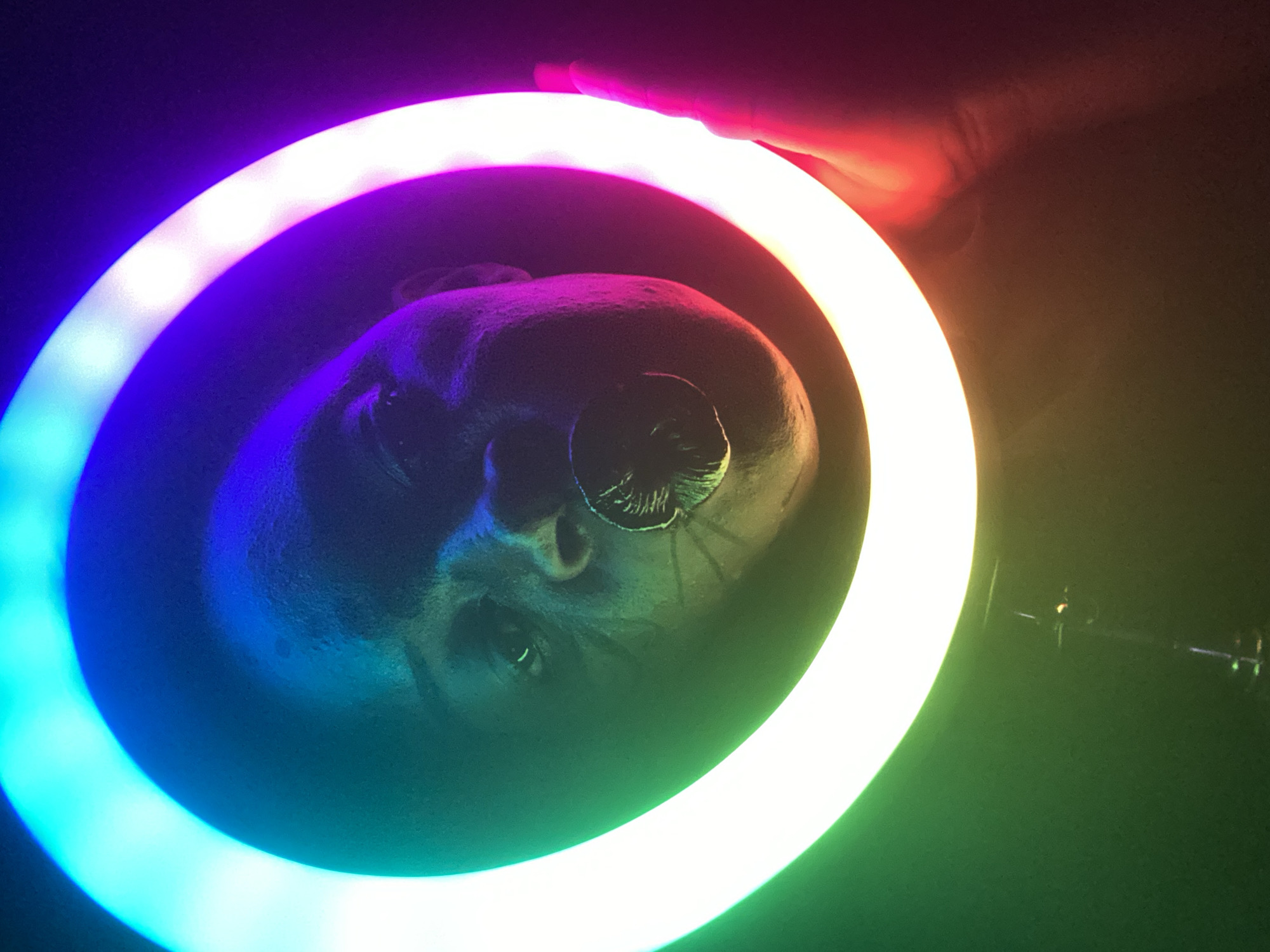 A person's face is presented within a colorful ring light. The person has dark skin and appears to be gazing upwards. The ring light transitions through various colors, including blue, purple, red, and green. A hand is visible at the top of the ring light, partially obscuring it. The environment is dark, with a stand partially visible at the bottom right. The location is not provided.