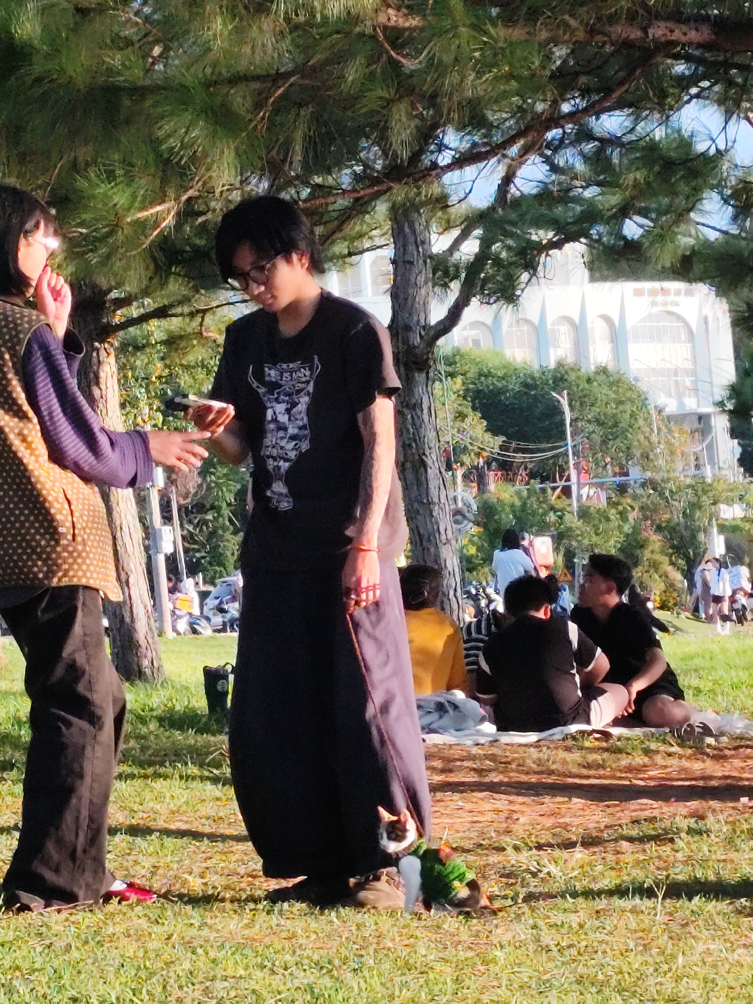 In the city of Da Lat, Vietnam, two individuals are visible in the foreground. The person on the left, wearing a patterned vest over a purple long-sleeved shirt and dark pants, is partially obscured. Their right hand is extended, and they appear to be holding a small object. The person on the right is wearing a black t-shirt with a graphic print and a long dark skirt or pants. They are looking down and holding a small device. Between their feet, a cat wearing a green vest is visible on the grass. In the middle ground, several other people are seated on a blanket on the grass, some wearing dark clothing. Behind them, a building with arched windows is visible through trees and foliage. The scene is set outdoors, on a grassy area with dappled sunlight filtering through trees, suggesting a park or public space.