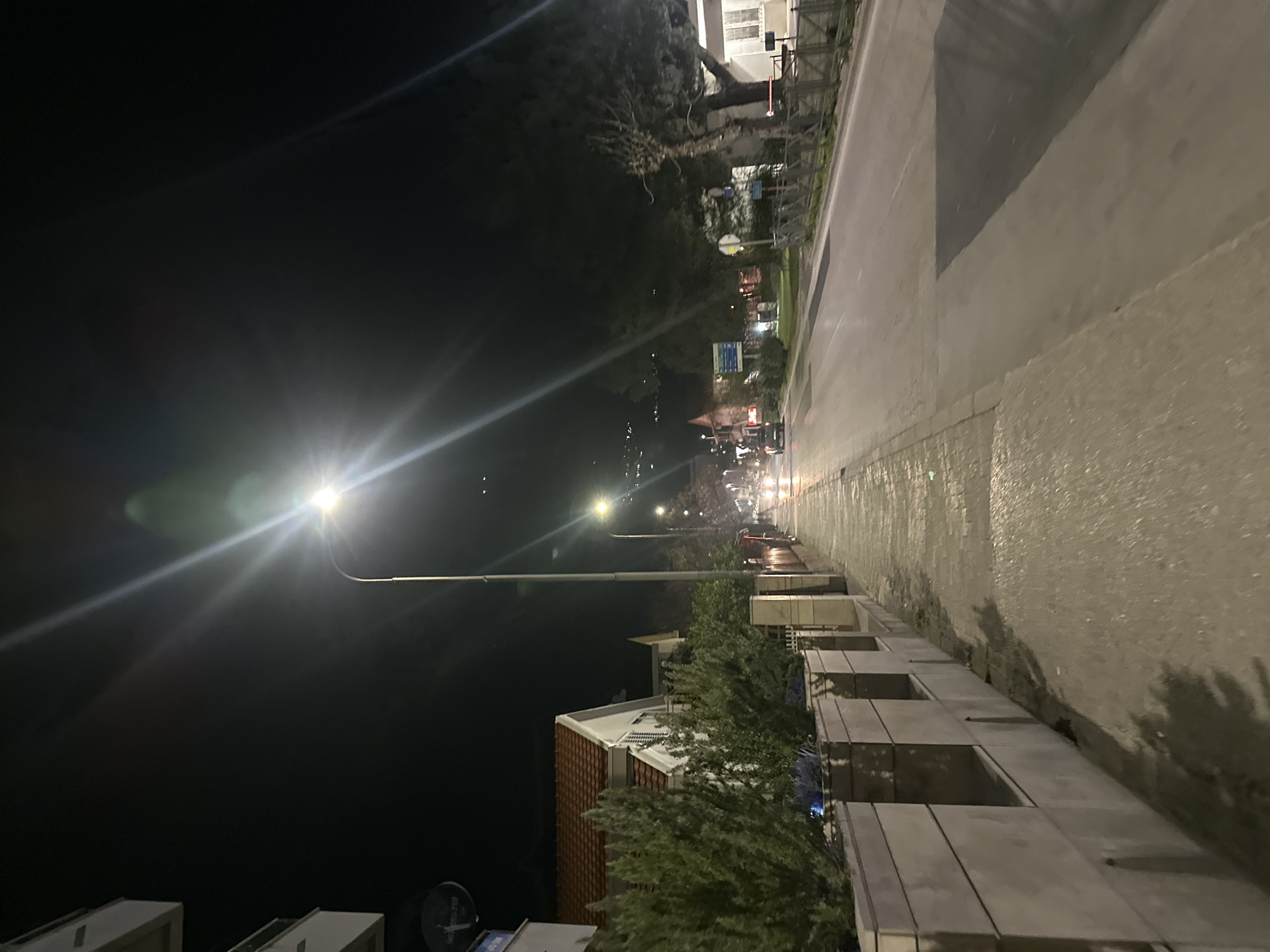 This image depicts a nighttime street scene in Budva, Montenegro. A paved sidewalk extends from the foreground along the left side of the frame, bordered by a concrete barrier featuring recessed sections and dark green shrubbery. Behind this, a residential building with a white facade and a red-tiled roof is visible, along with a satellite dish mounted on its exterior. Two bright streetlights illuminate the scene, casting significant lens flares, with one streetlight positioned centrally in the mid-ground. To the right of the sidewalk is an asphalt road. Multiple vehicles with illuminated headlights and taillights are present in the distance on the road, indicating traffic flow. The right side of the street features trees, a metal fence, and distant structures with lights. The background shows a dark sky with scattered lights, suggesting distant buildings or terrain elevation. No individuals are visible on the sidewalk or road.