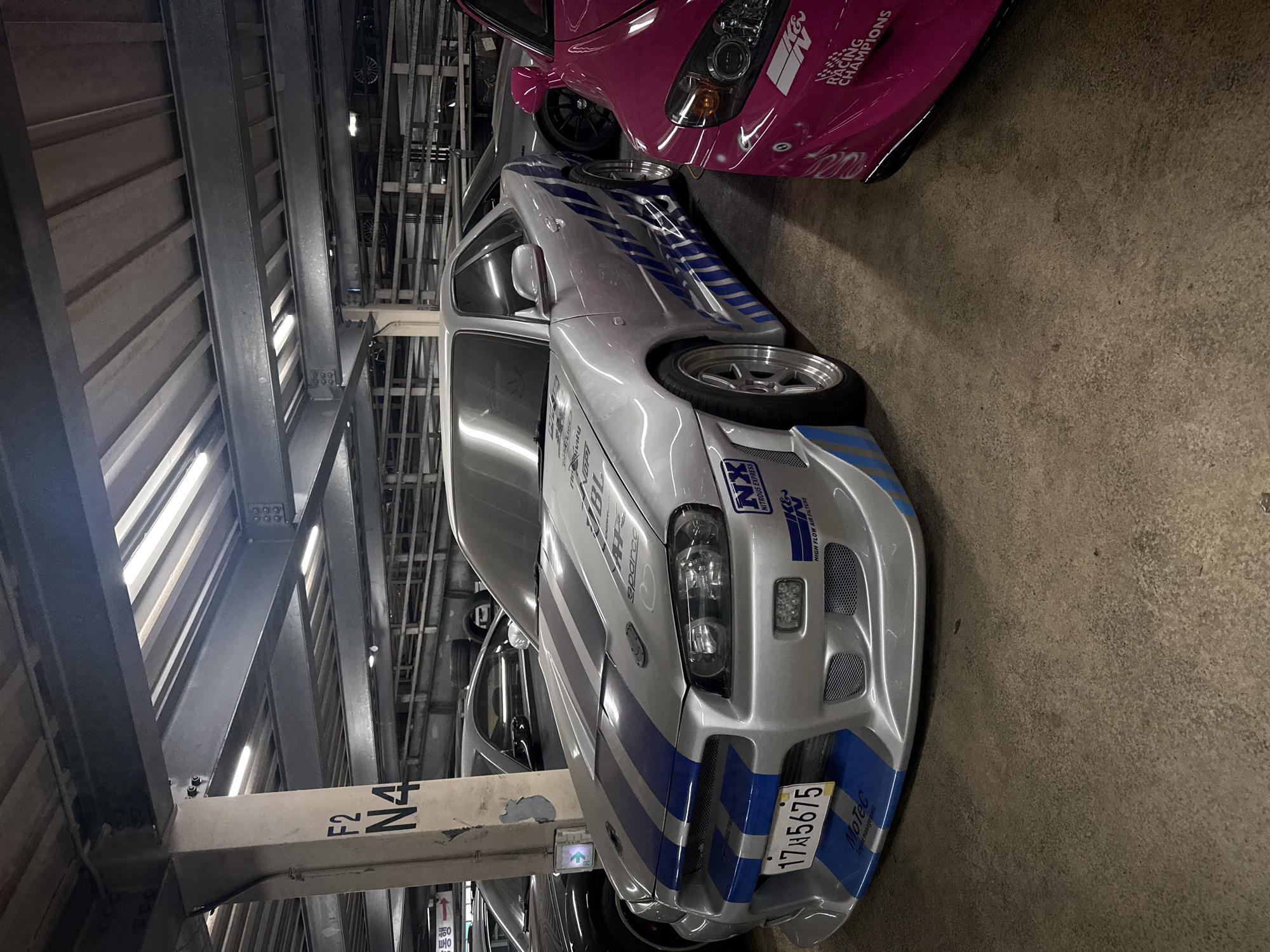 The image shows a silver Nissan Skyline R34 with blue racing stripes parked in a multi-story parking garage. The car has a license plate that reads "17서5675". The car has a sticker on the front that reads "NX NITROUS EXPRESS" and "HIGH FLOW AIR FILTERS". There is a pink car parked next to the silver car, with a sticker on the side that reads "RACING CHAMPIONS". The parking garage is located in the city of Seoul, South Korea, based on the license plate and the Korean text on the column. The column is labeled "F2 N4".