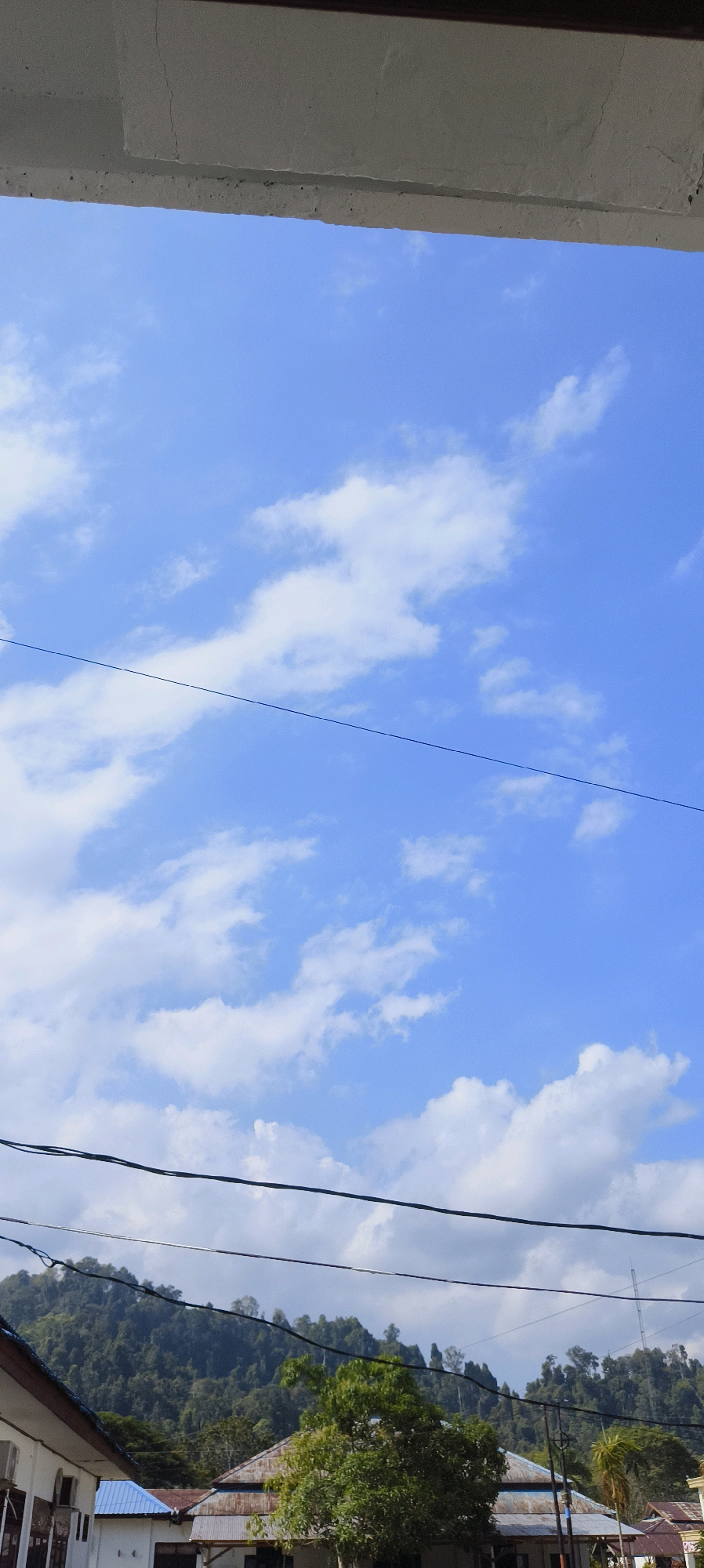The image captures a clear blue sky dotted with fluffy white clouds. In the foreground, a concrete overhang is visible at the top of the frame, with electrical wires stretching horizontally across the sky. Below the sky, a lush green hillside forms the backdrop, covered in dense trees. In the lower part of the image, several buildings with various roof types, including corrugated metal and blue-tiled roofs, are partially visible. The presence of electrical wires and buildings suggests a populated area, possibly within the city of Waisai, Indonesia. The overall scene is bright and suggests a daytime setting. There are no people or animals visible in the frame.