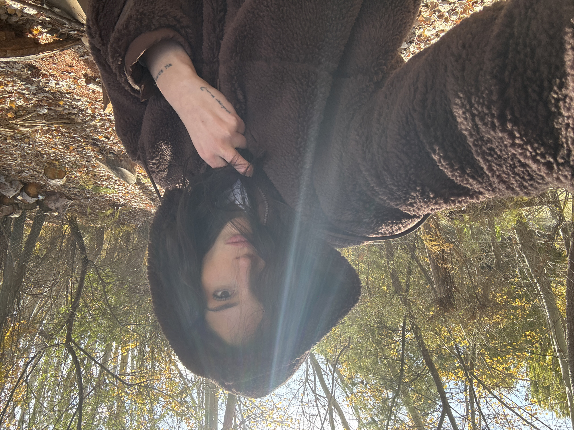 A selfie image shows a person wearing a brown hooded jacket against a backdrop of trees, presumably outdoors. The trees suggest a wooded area, possibly a park or forest. The lighting indicates daytime, with some sun glare. The person's face is partially obscured by their hair and the hood. Visible tattoos are on one wrist. The image does not provide explicit details that would allow for location in the city of Trelew, Argentina, or any other specific geographical location.