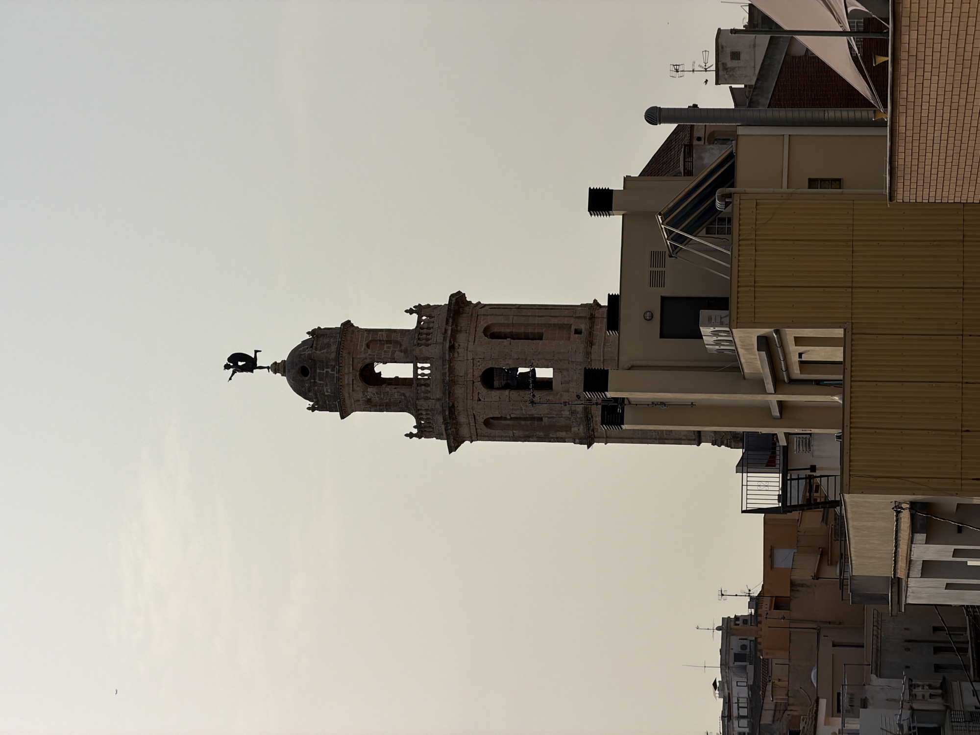 The image shows a view of buildings in the city of El Vendrell, Spain, with a prominent bell tower of a church in the center. The sky is overcast and pale. The bell tower is made of stone and has a statue of a figure on top. Several other buildings, varying in height and architectural style, surround the bell tower. Some of these buildings have rooftop features such as antennas, chimneys, and air conditioning units. The overall scene depicts an urban landscape with a mix of historic and modern structures under a diffused light.