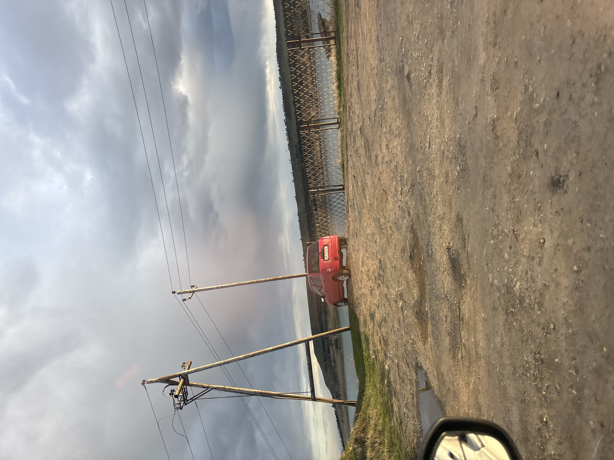 The image shows a dirt road in Tbilisi, Georgia, with a red car parked on the side. A fence runs parallel to the road, and a body of water is visible beyond the fence. Overhead power lines stretch across the sky, which is filled with grey clouds. The image appears to be taken from inside a vehicle, as a side mirror is visible.