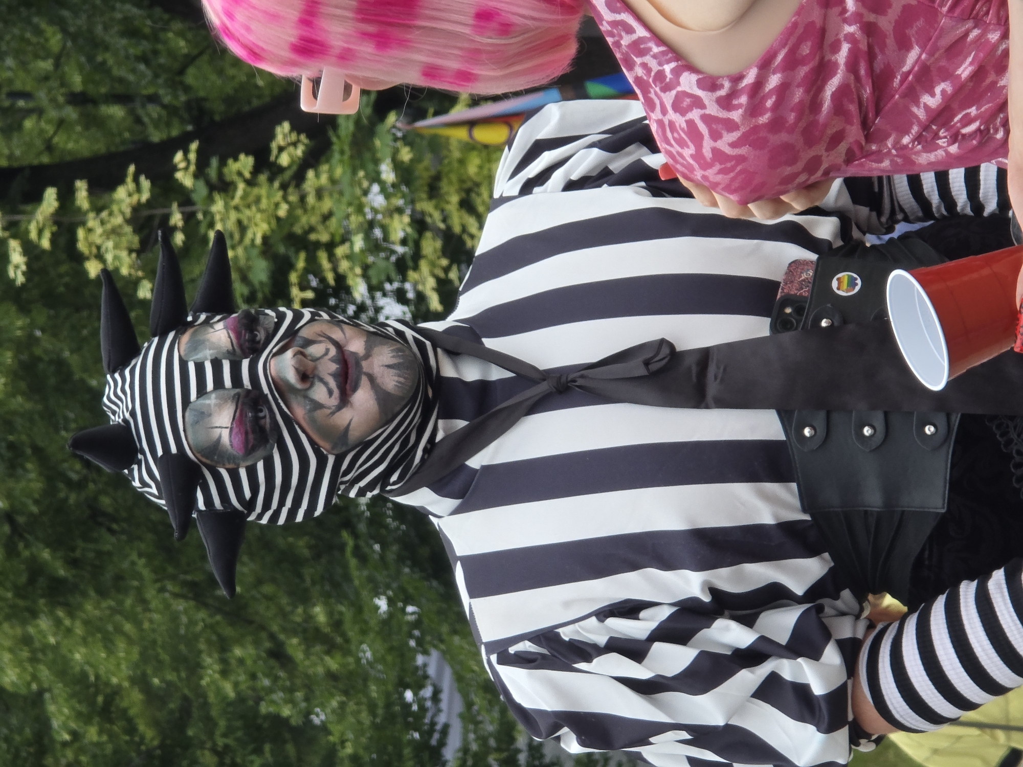 The image shows a person in a black and white striped outfit with a spiked headpiece and elaborate face paint. The person is wearing a black tie. Another person with bright pink hair is visible on the right side of the frame, wearing a pink leopard print dress. A red plastic cup is held by the person on the right. In the background, there are trees and greenery, suggesting an outdoor setting. There is a pride flag sticker visible on a phone case. The scene appears to be at a public event, possibly a parade or festival. This photo was taken on June 18, 2023, in Warsaw, Poland, during the annual Equality Parade.