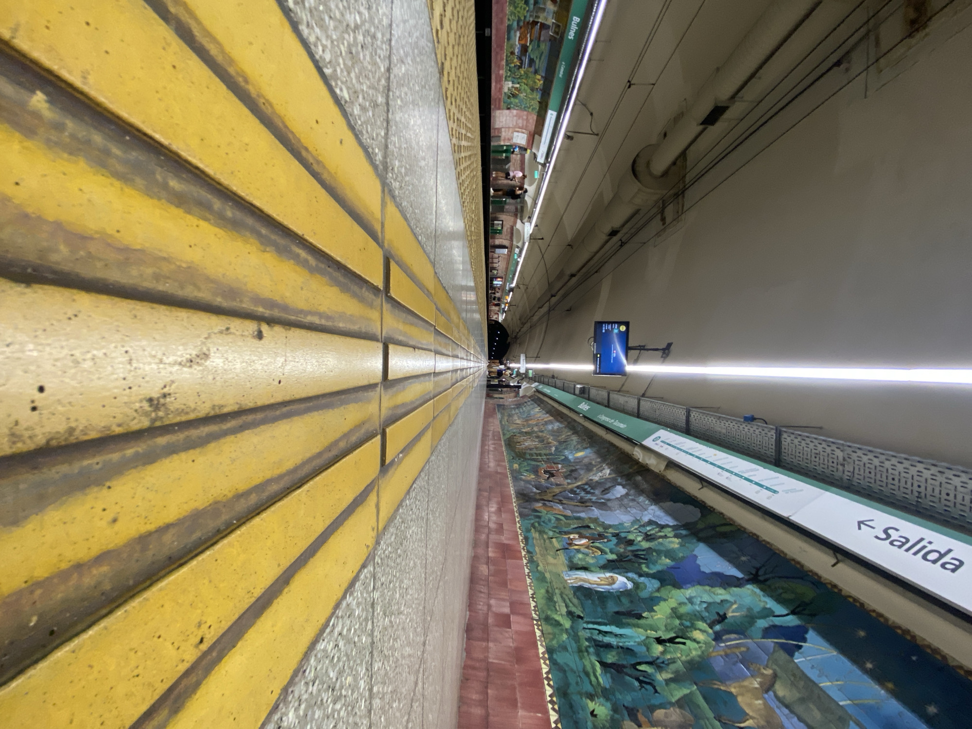 The image shows the Bulnes subway station in Buenos Aires, Argentina. The camera is positioned on the platform, looking down the line toward the end of the tunnel. On the left is the edge of the platform marked with yellow tactile tiles. A tiled wall is also on the left. Along the right side of the frame, a long mural extends along the station wall above a sign with the word "Salida" (exit) and an arrow indicating the direction of the exit. Toward the end of the platform, there are several people waiting. Overhead lighting is visible along the station.