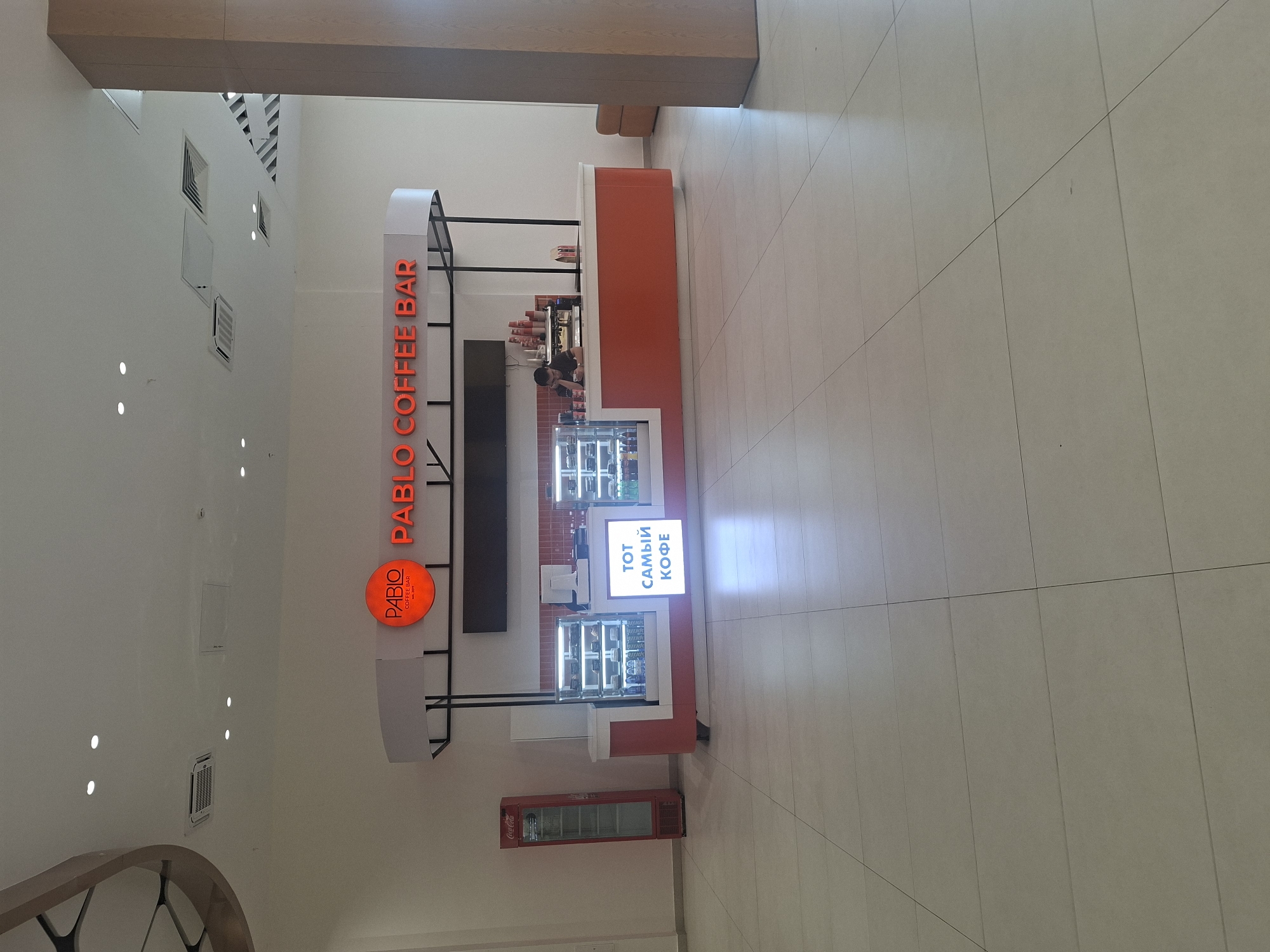 The image shows a "Pablo Coffee Bar" kiosk in what appears to be a shopping mall in Kokshetau, Kazakhstan. The kiosk is brightly lit with a white and orange color scheme and has the name "PABLO COFFEE BAR" vertically displayed in orange lettering. There's also a smaller round sign with the Pablo logo. A person is visible behind the counter, likely a barista. The kiosk has refrigerated display cases filled with drinks and food items. A Coca-Cola refrigerator is located next to the kiosk. The floor is tiled. The mall's interior is illuminated by recessed ceiling lights and appears spacious. A support beam is visible in the upper portion of the image.