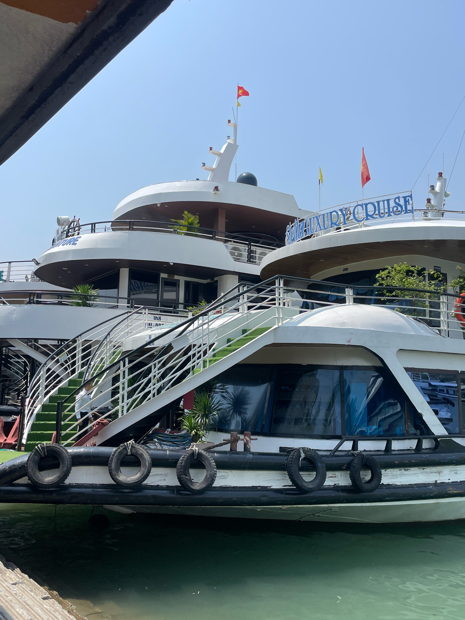 A white luxury cruise ship is docked in the water in Quang Ninh City, Vietnam. The ship has multiple decks, white railings, and blue lettering that spells out "LUXURY CRUISE". Green carpet runners are laid on outdoor staircases that lead up to the upper decks. Several black tires are attached to the side of the ship as fenders. The water around the ship is a murky green color. The sky above is clear and blue.