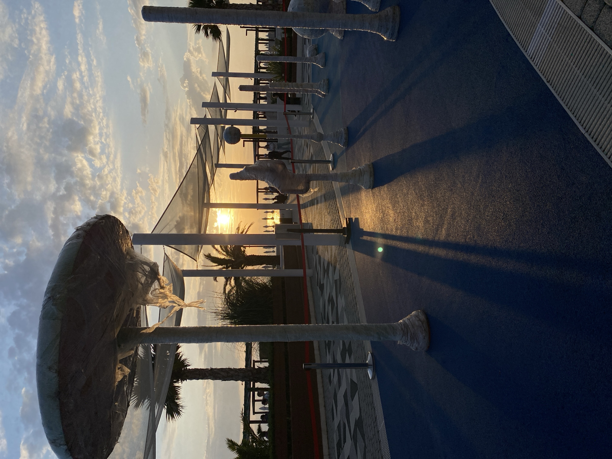The image shows an outdoor scene in Sochi, Russia, at sunset. In the foreground are several covered sculptures, likely awaiting unveiling, placed on a blue surface. Behind these, a row of modern canopy structures provides shade. The sunset is visible through the structures, with palm trees and other foliage in the background. The image is taken from a low angle, with a slight upward tilt, and the scene appears calm and serene. No people are immediately visible.